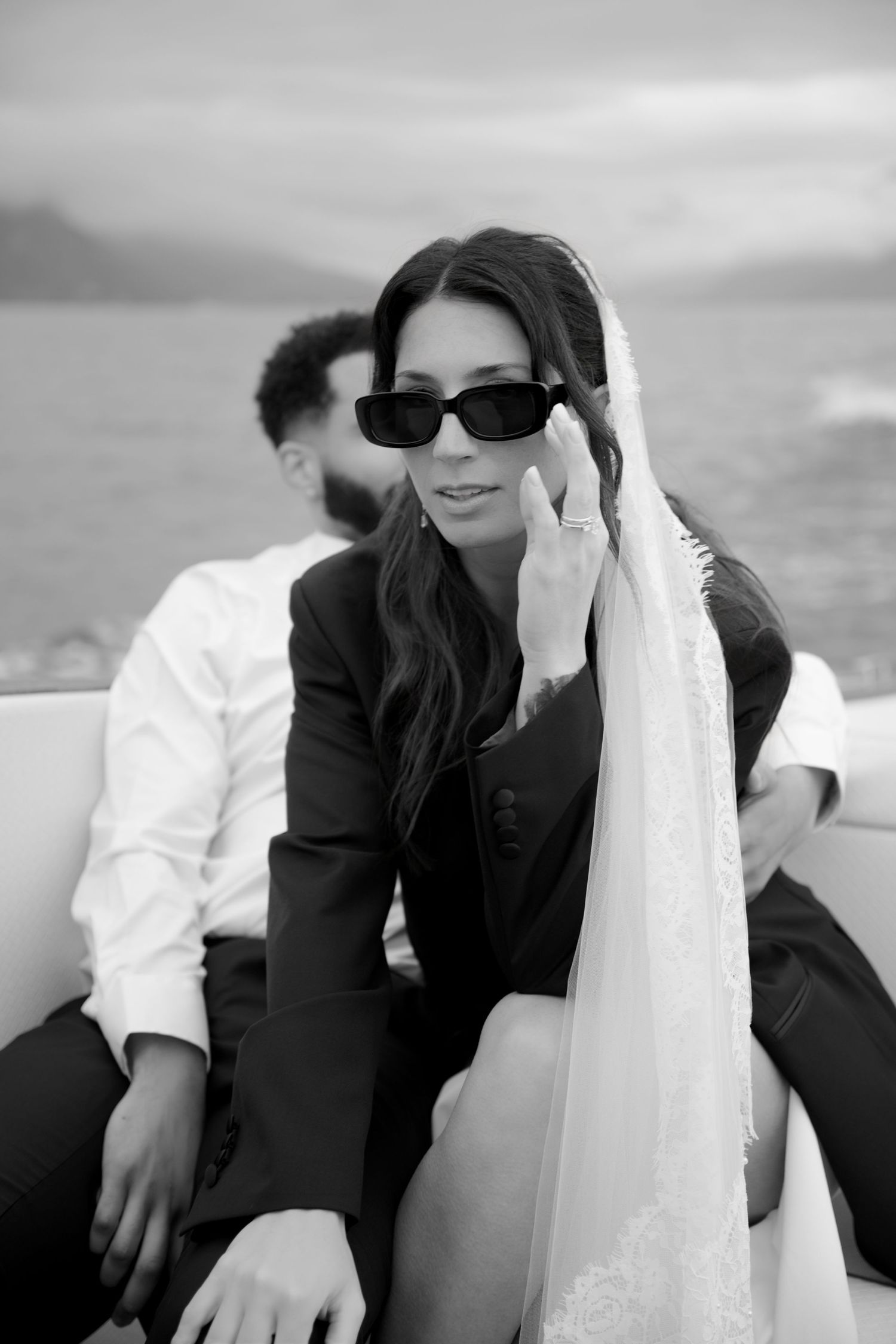 Black and white photo of a bride in sunglasses holding her veil on a boat with groom seated behind her.