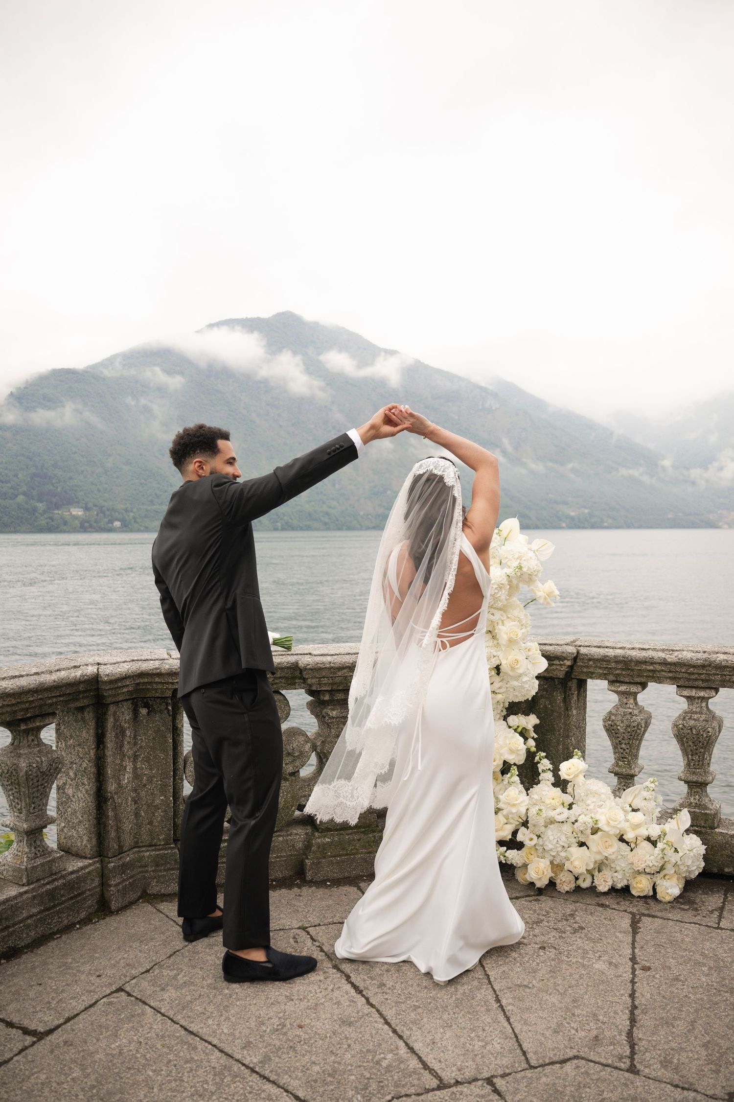Groom twirls bride in white gown and veil on elegant stone balcony overlooking misty mountain lake in Italy.