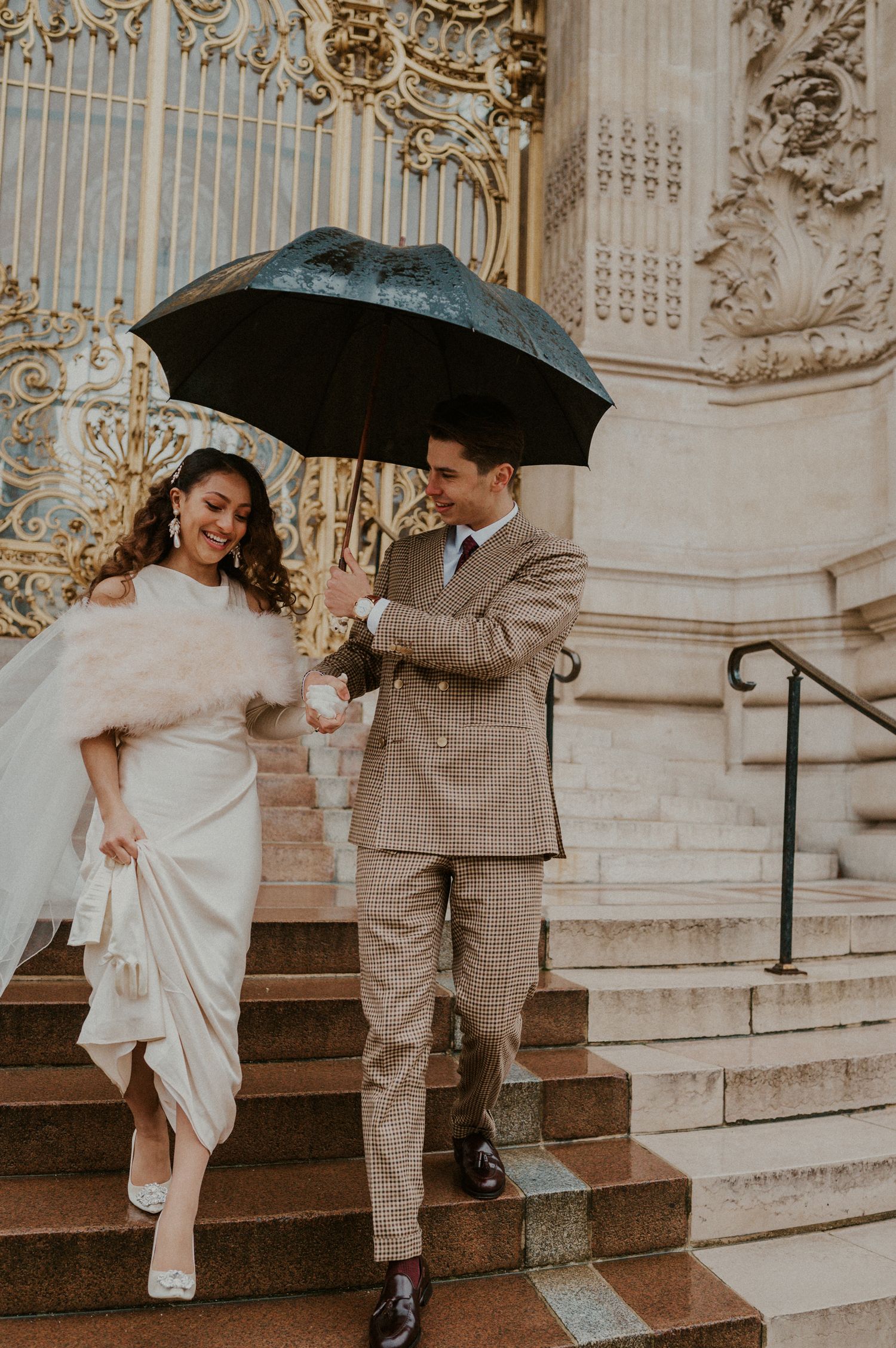 Classic wedding couple descending ornate marble steps with a black umbrella under elegant architectural details.