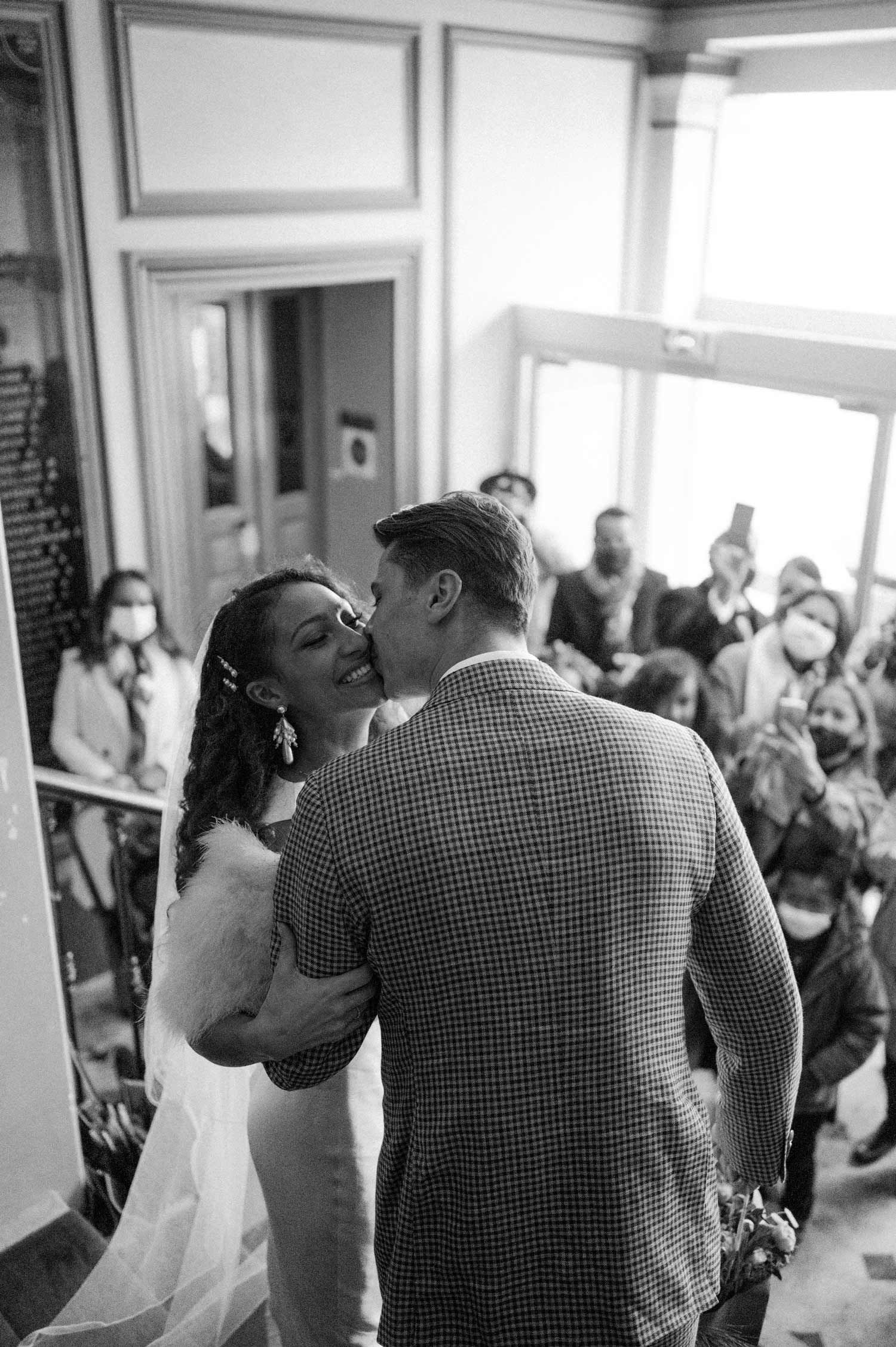 A black and white wedding photo capturing a kiss with guests watching in the background.