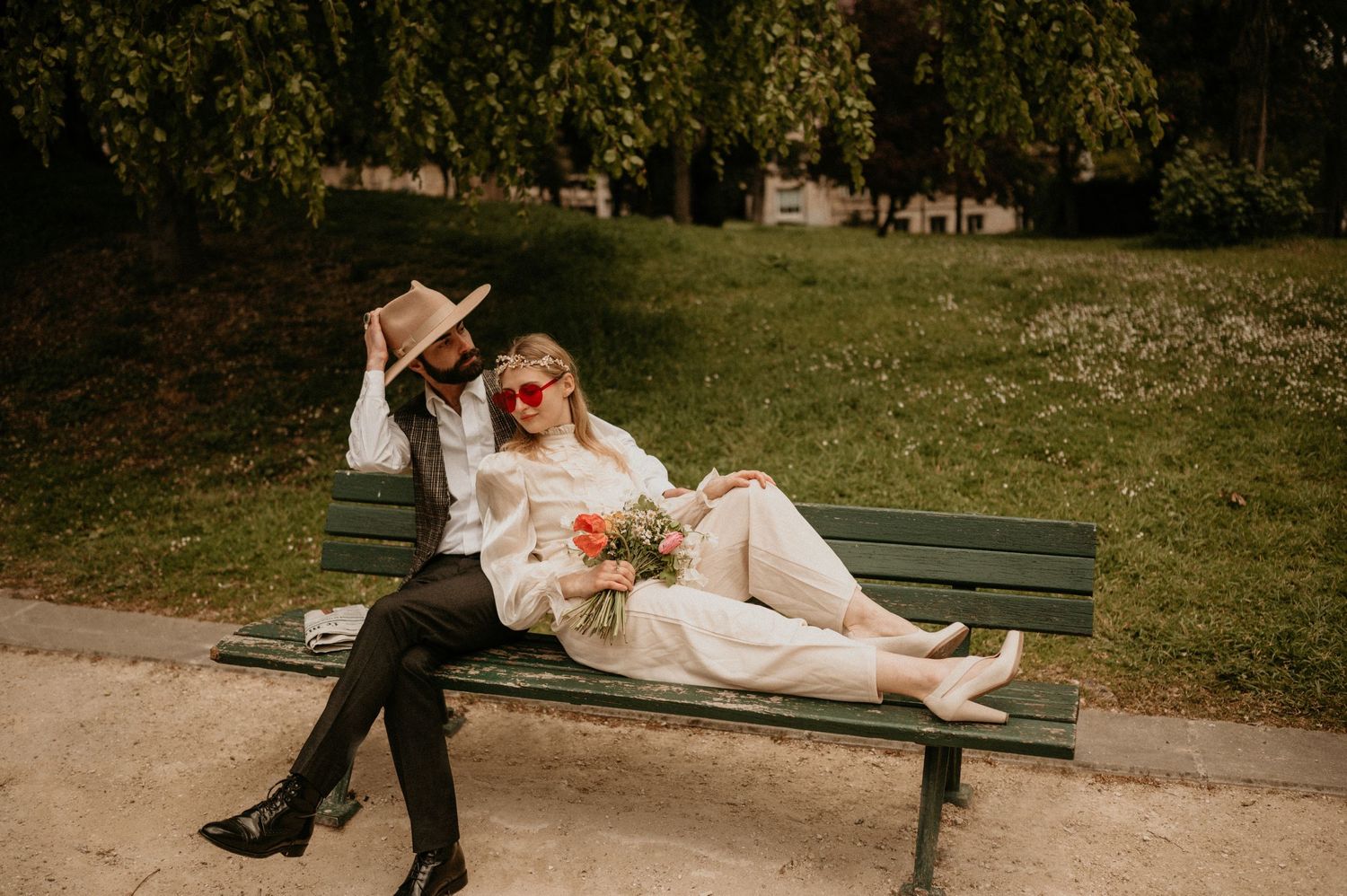 A couple sits casually on a wooden bench in a park setting with dreamy vintage styling.