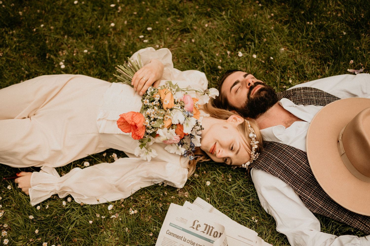A couple lying on grass with flowers and a newspaper, captured in a romantic vintage-style photo.