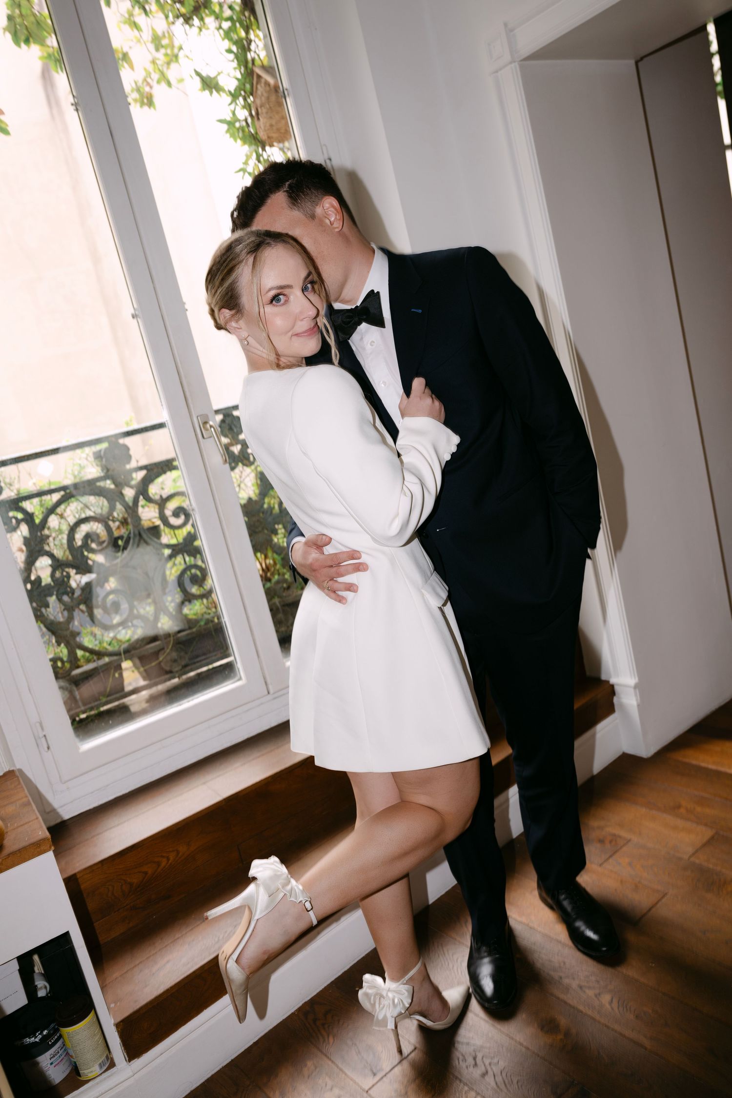 Romantic couple embraces near a French window, with her wearing a white mini dress and him in classic black tie.