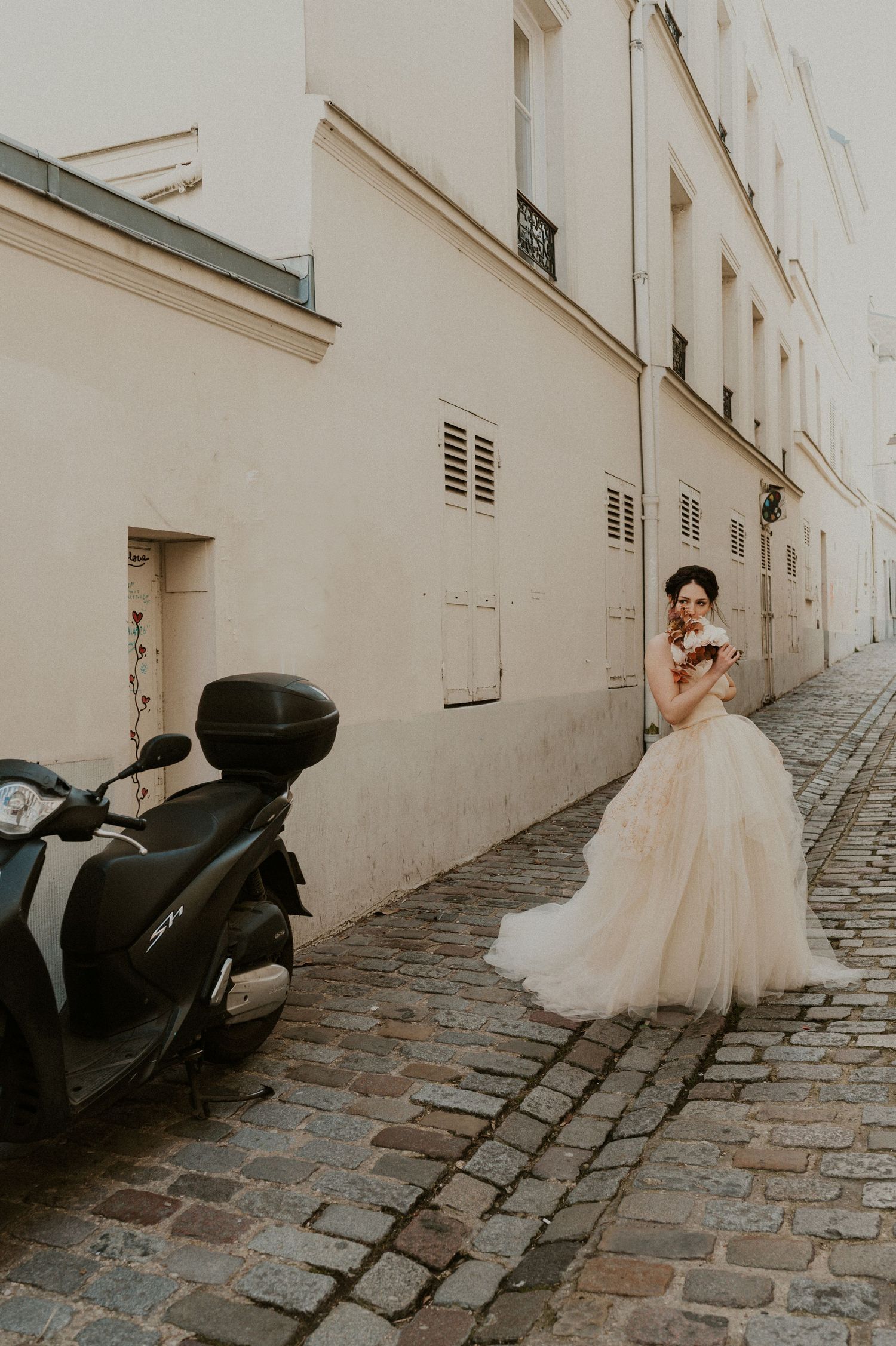Bride in champagne gown poses beside scooter on narrow Parisian alleyway.