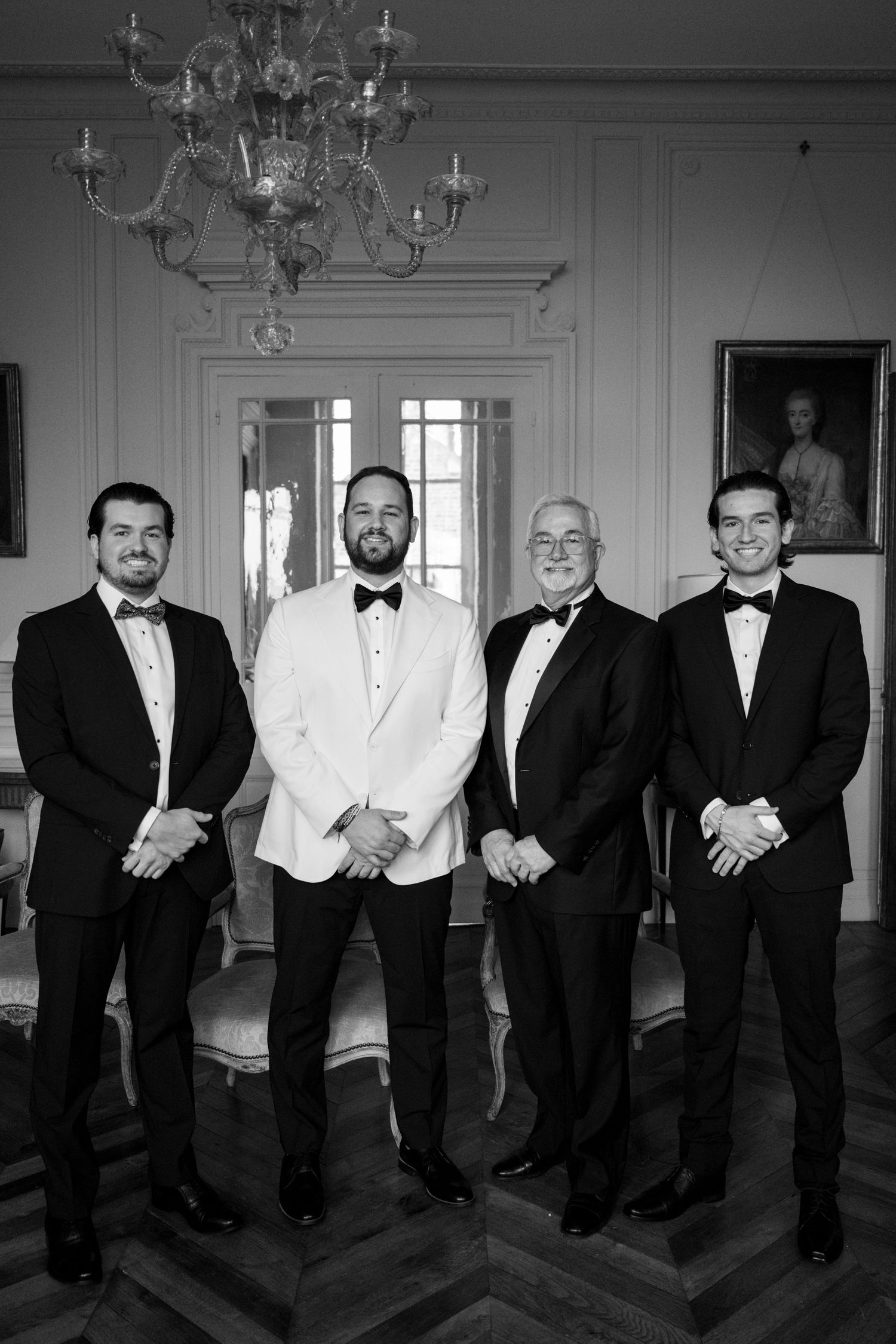 Groomsmen in formal black and white tuxedos pose together in an elegant room with chandeliers and classic decor.