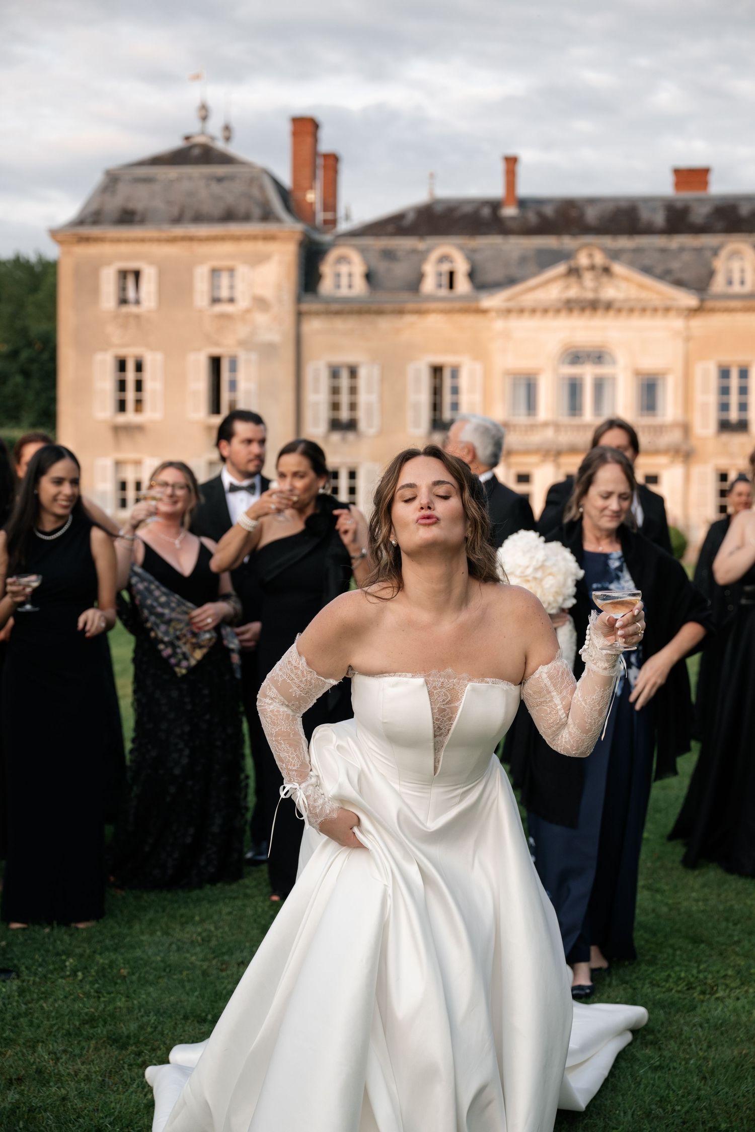 Bride in strapless white gown poses confidently before French chateau with guests in black attire.