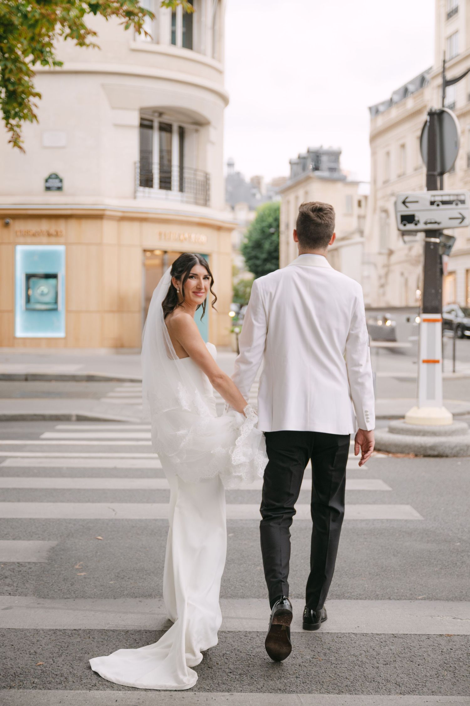 Bride in white gown and groom in white jacket hold hands while crossing a Parisian street.