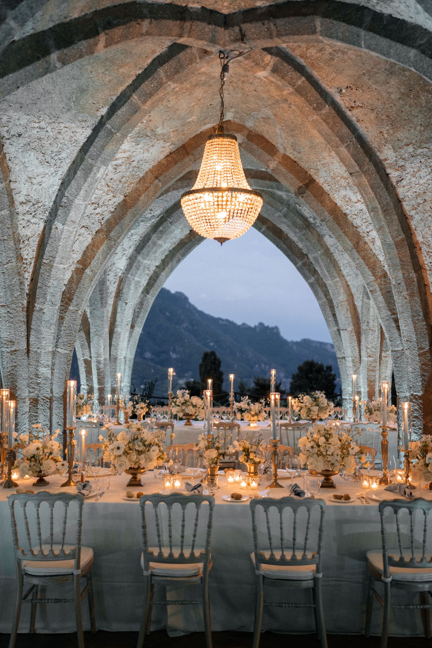 Stunning wedding reception setup under Gothic stone arches with chandelier, candlelit tables, and mountain views.