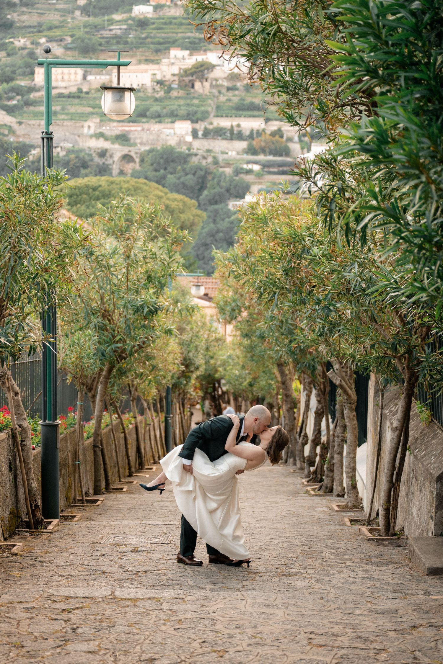 Bride and groom sharing a romantic dip kiss on a tree-lined cobblestone path in Italy.