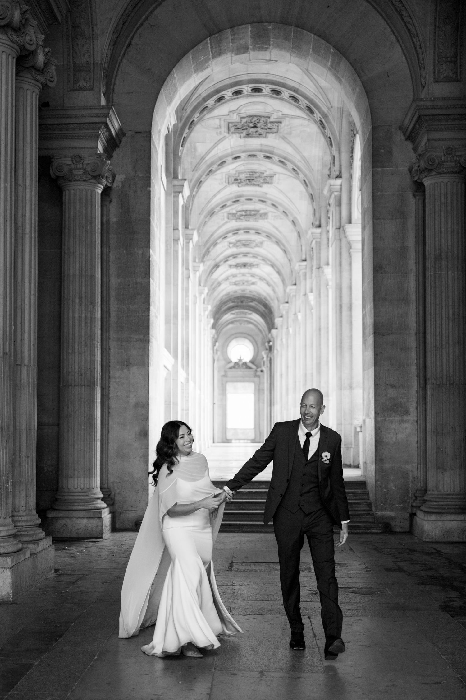 Black and white photo of wedding couple walking through grand arched colonnade at the Louvre.