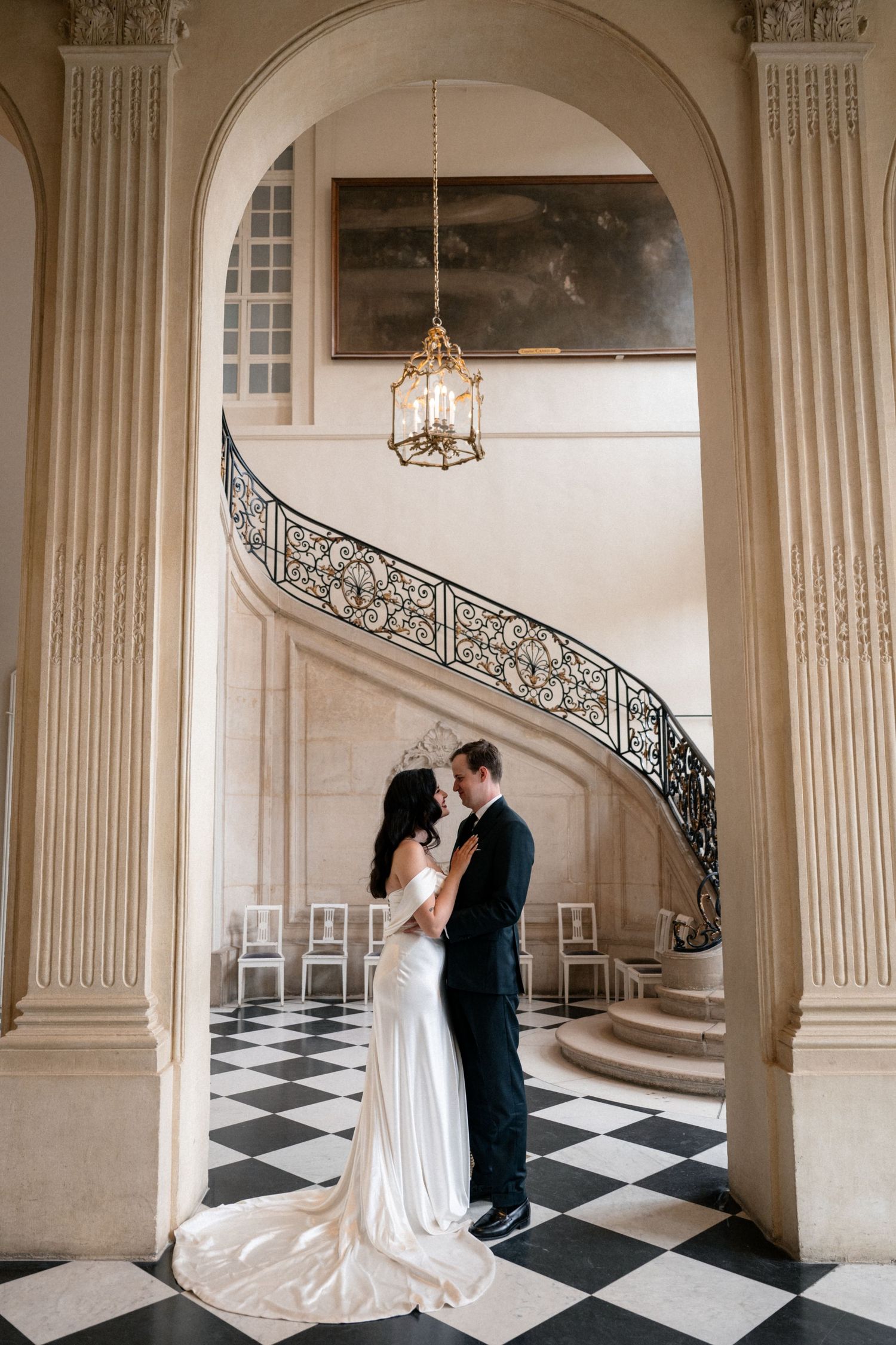 Bride and groom share intimate moment under grand arch with ornate spiral staircase and checkered marble floor.