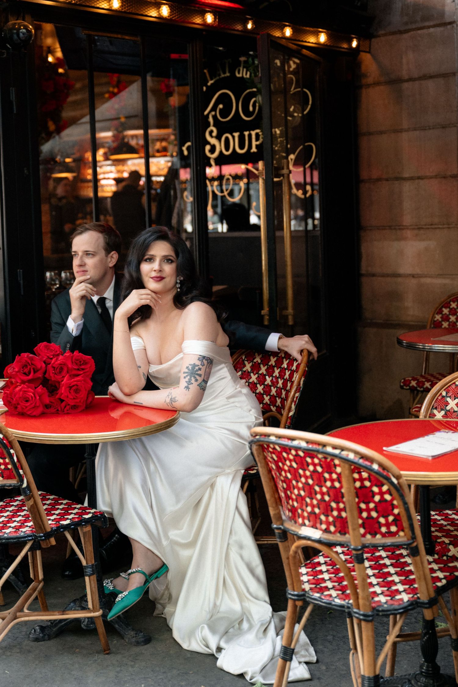 Bride and groom seated at classic red Parisian cafe terrace with bouquet of red roses on table.