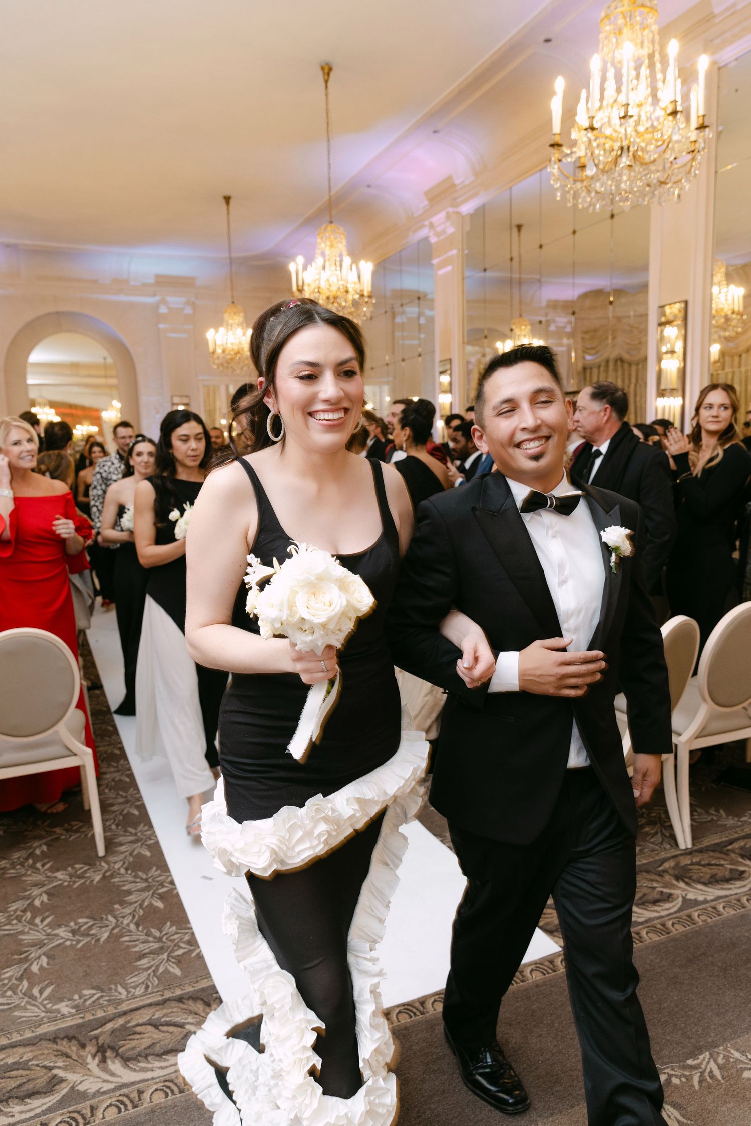 Smiling bridesmaid in black gown holding white bouquet walks arm-in-arm with groomsman in tuxedo at elegant wedding reception.