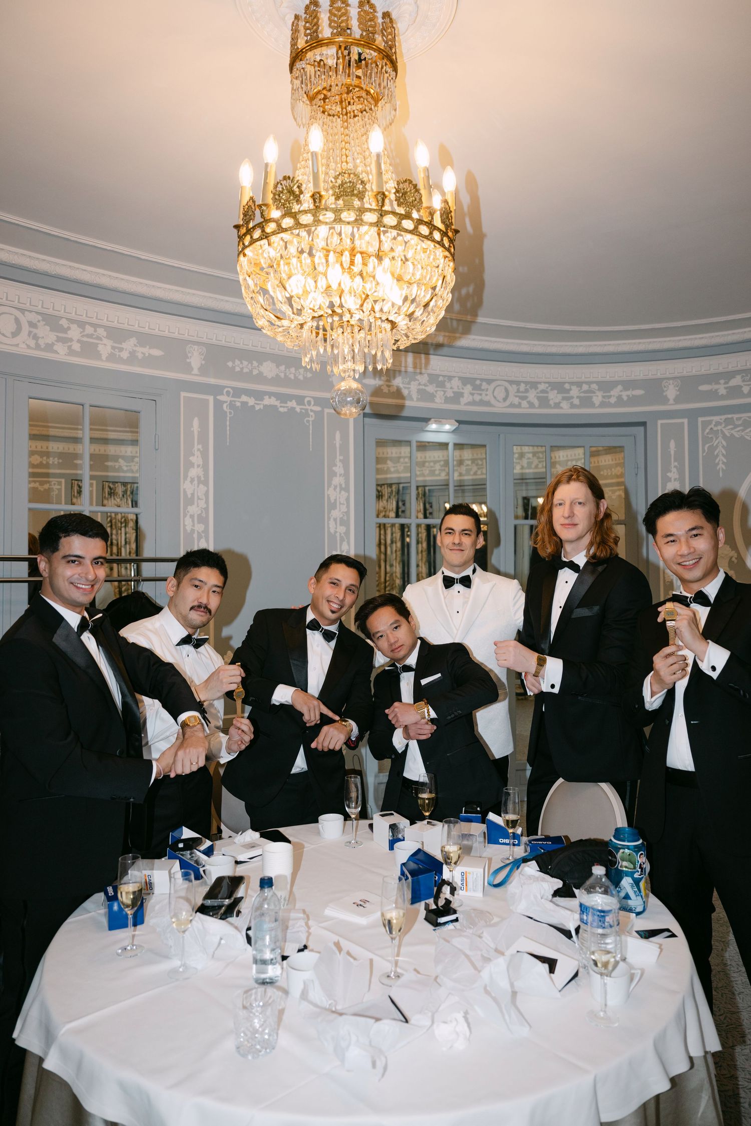 Groomsmen in black tuxedos posing together under crystal chandelier in elegant blue reception room.