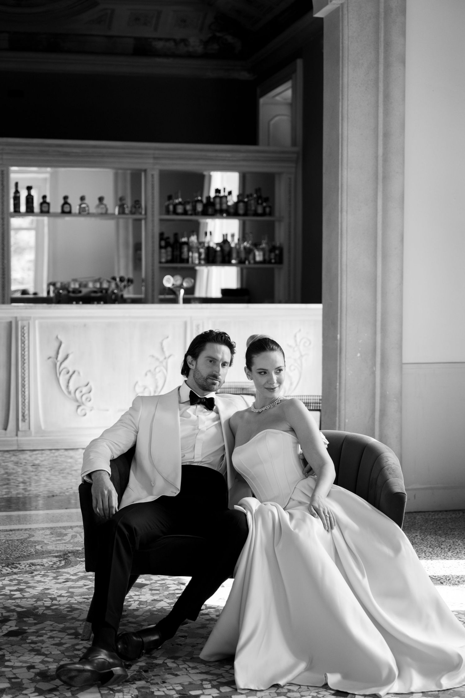 Black and white portrait of stylish bride and groom seated in luxury Italian hotel bar.
