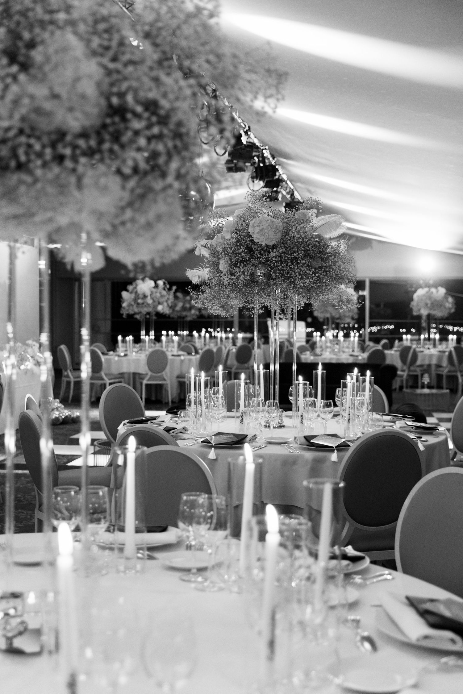 Black and white photo of elegant wedding reception tables with tall floral centerpieces and candles.