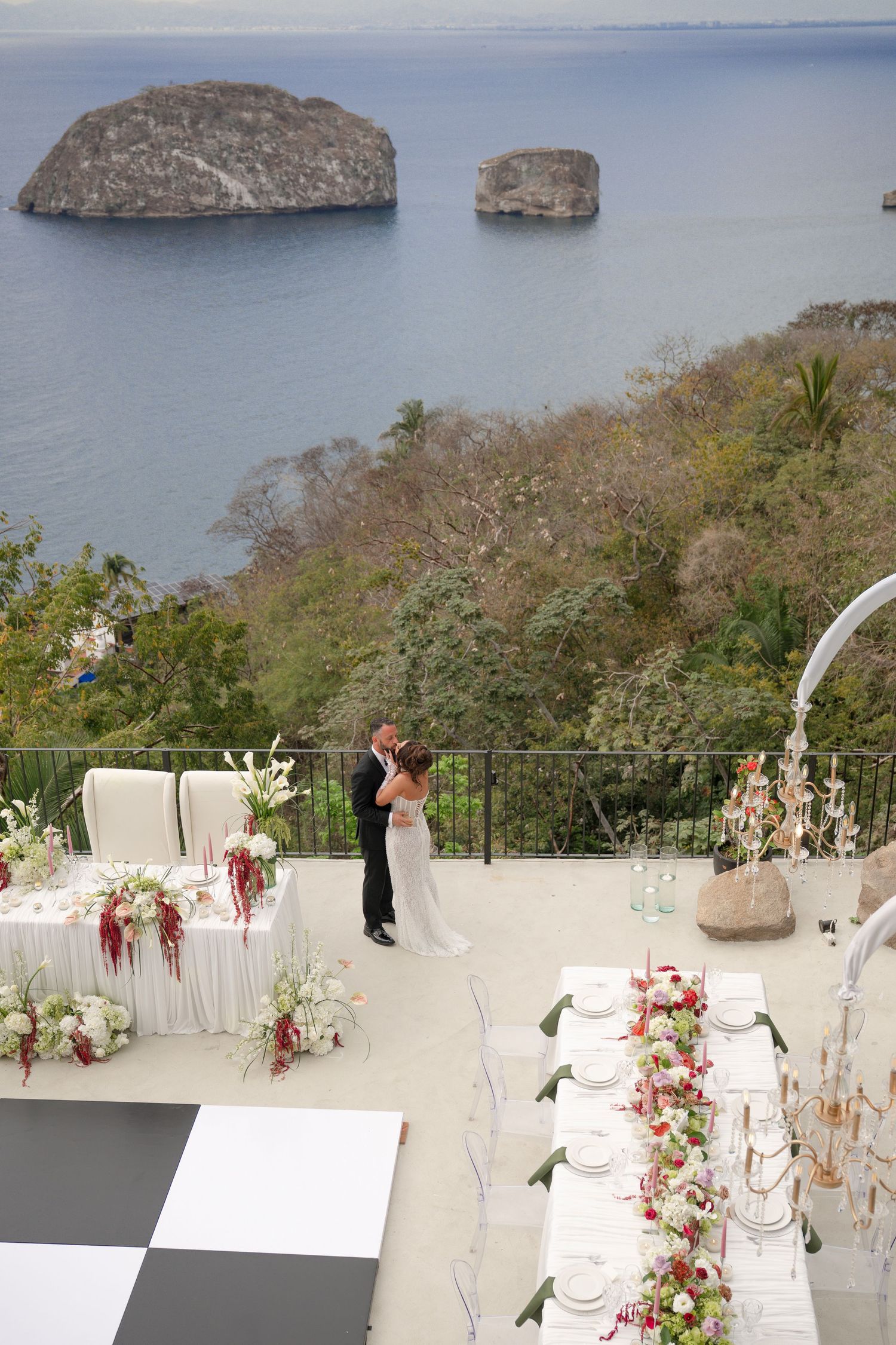 Bride and groom embrace on a cliffside terrace with ocean island views and elegant floral reception tables.