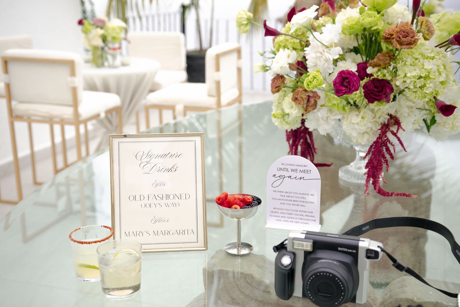 Elegant wedding reception table with floral arrangements, signature cocktail signs, and a Fujifilm Instax camera on a glass surface.