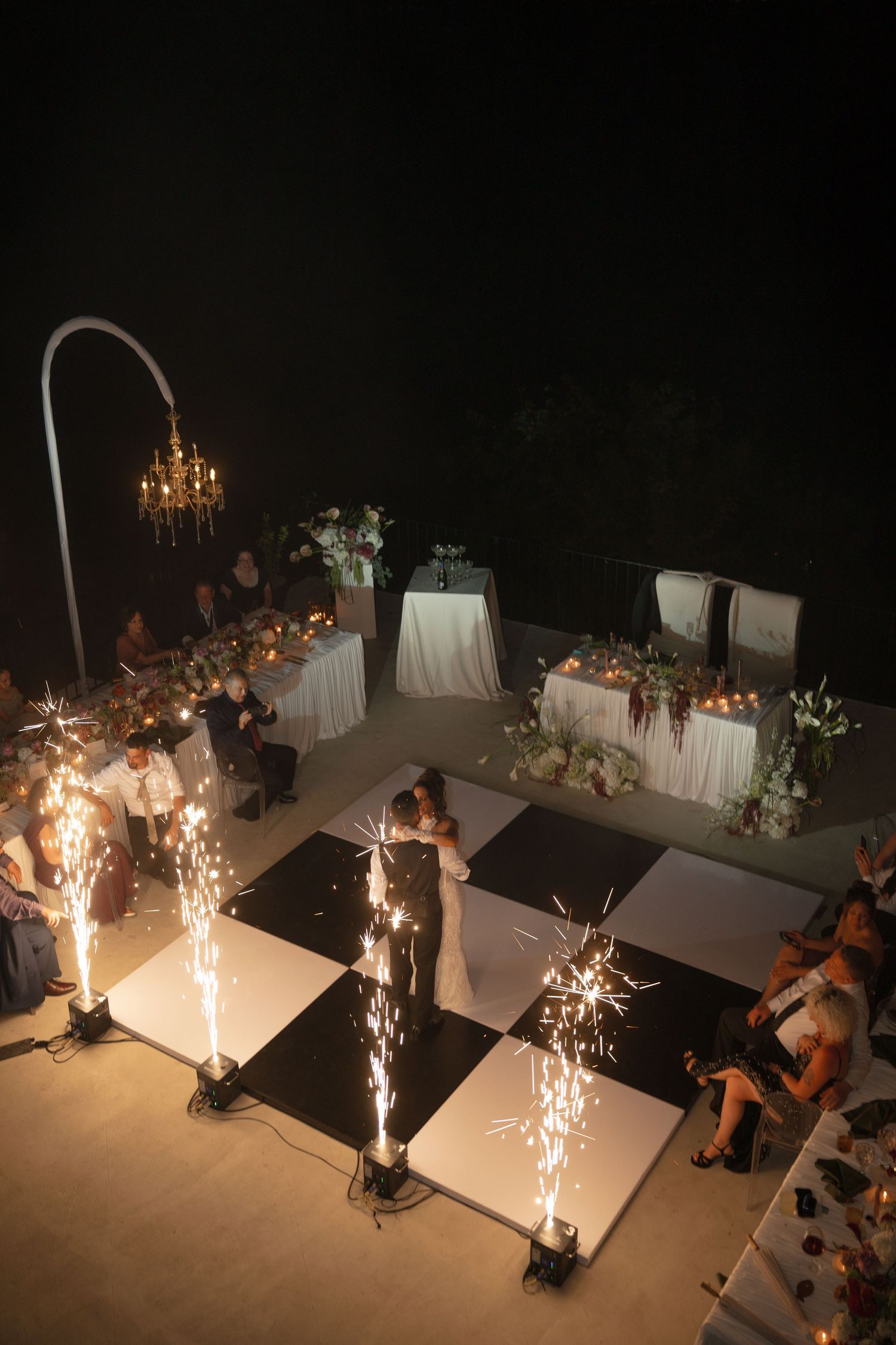Aerial view of an elegant outdoor wedding reception with a black and white checkered dance floor and sparkling cold fireworks.