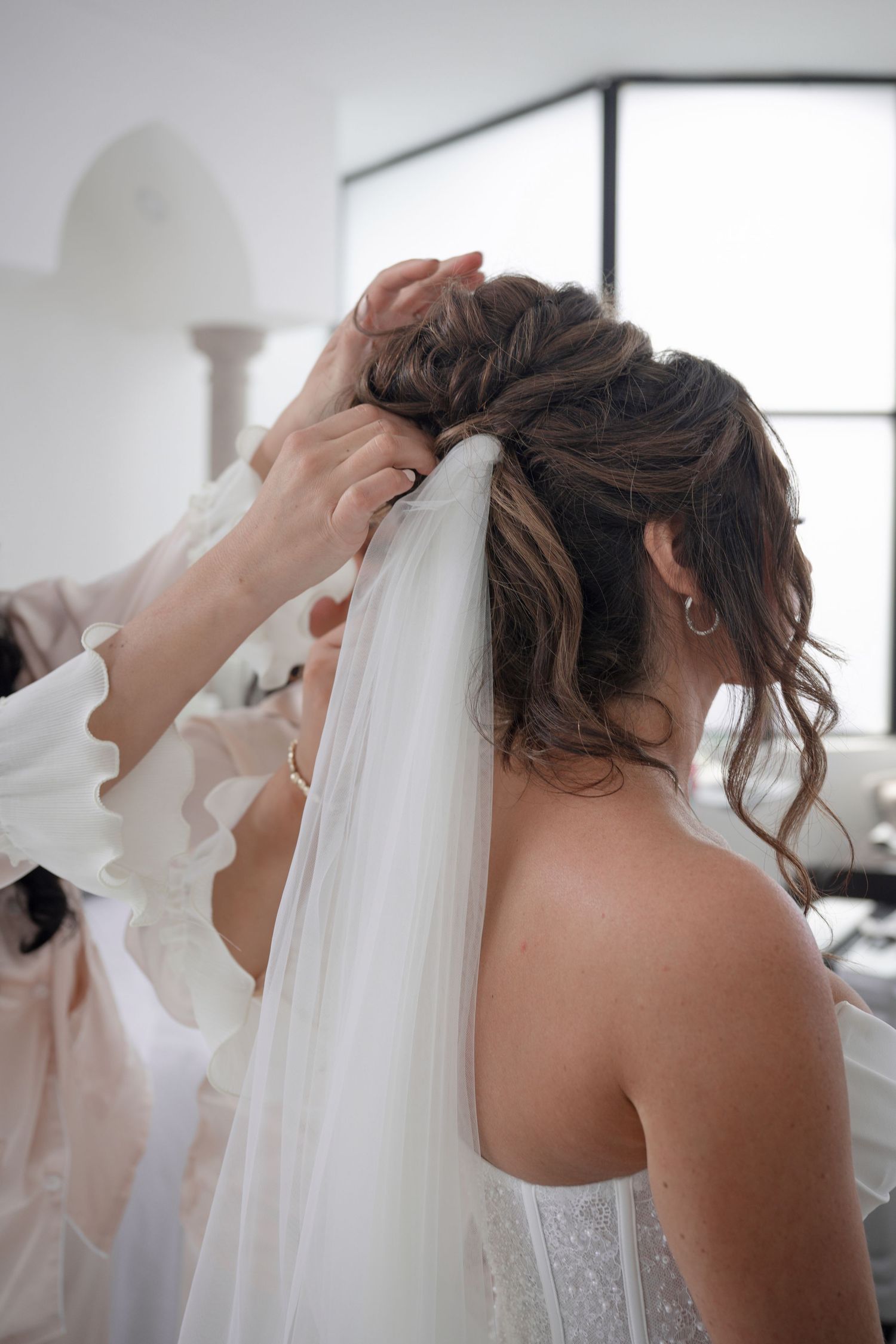Hairstylist carefully attaches a flowing white veil to the bride's elegant updo hairstyle before the wedding.