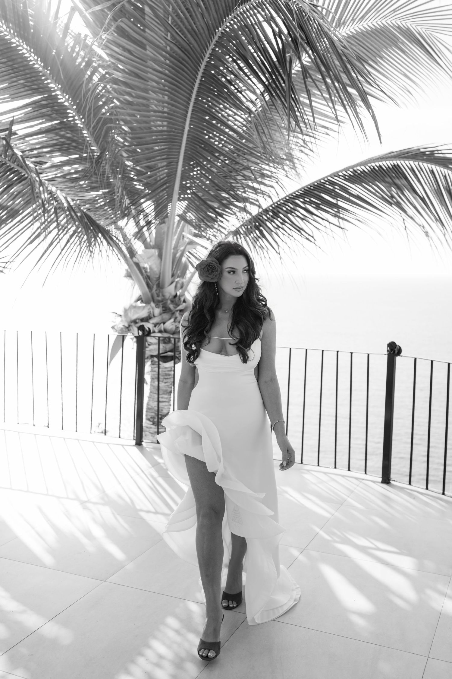 Black and white photo of a woman in a flowing white dress standing on a tropical terrace with palm trees and ocean view.