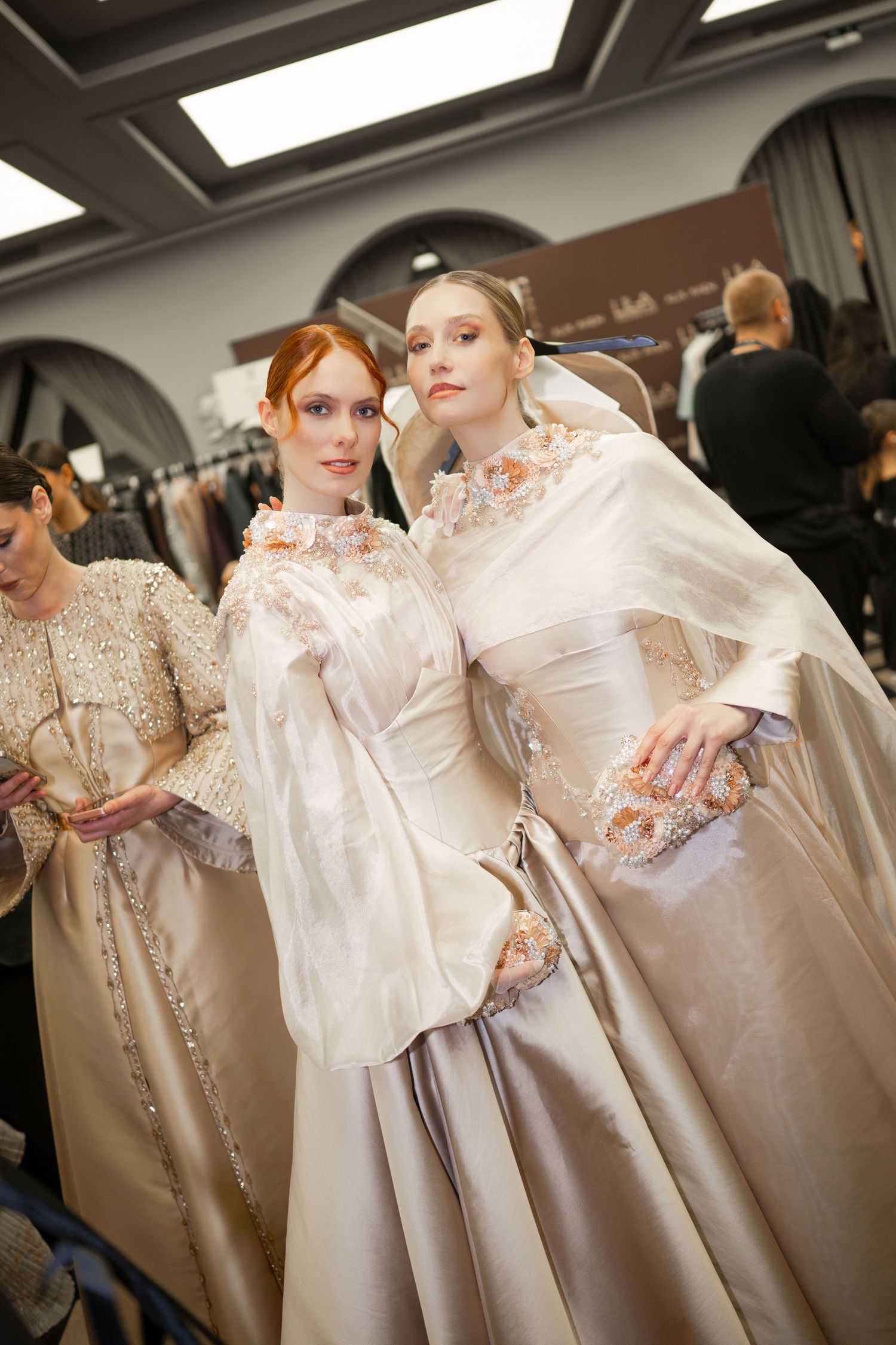 Two models pose backstage in elegant ivory and silver ball gowns with floral embellishments at a fashion show.
