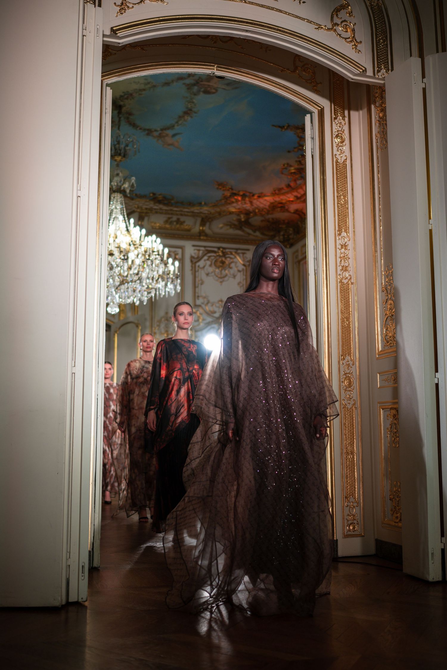 Models walk in ornate gilded hall wearing flowing embellished gowns at a luxury fashion show.