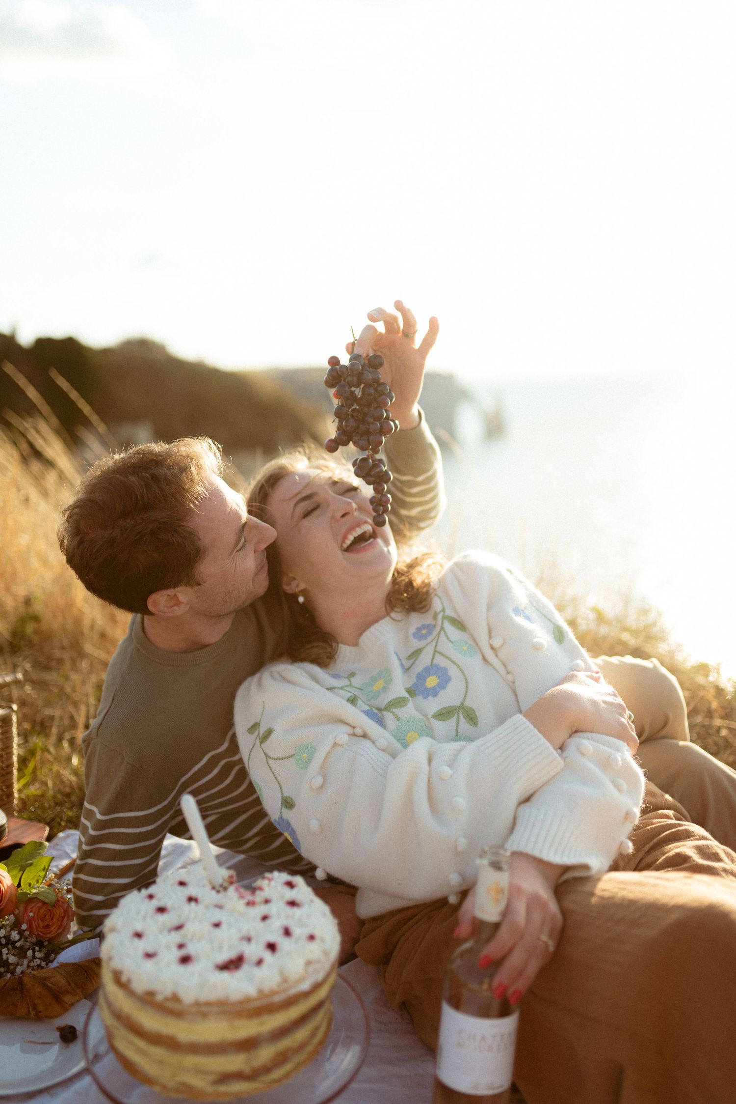 Happy couple enjoys a clifftop picnic with cake and grapes, laughing together in golden sunlight.
