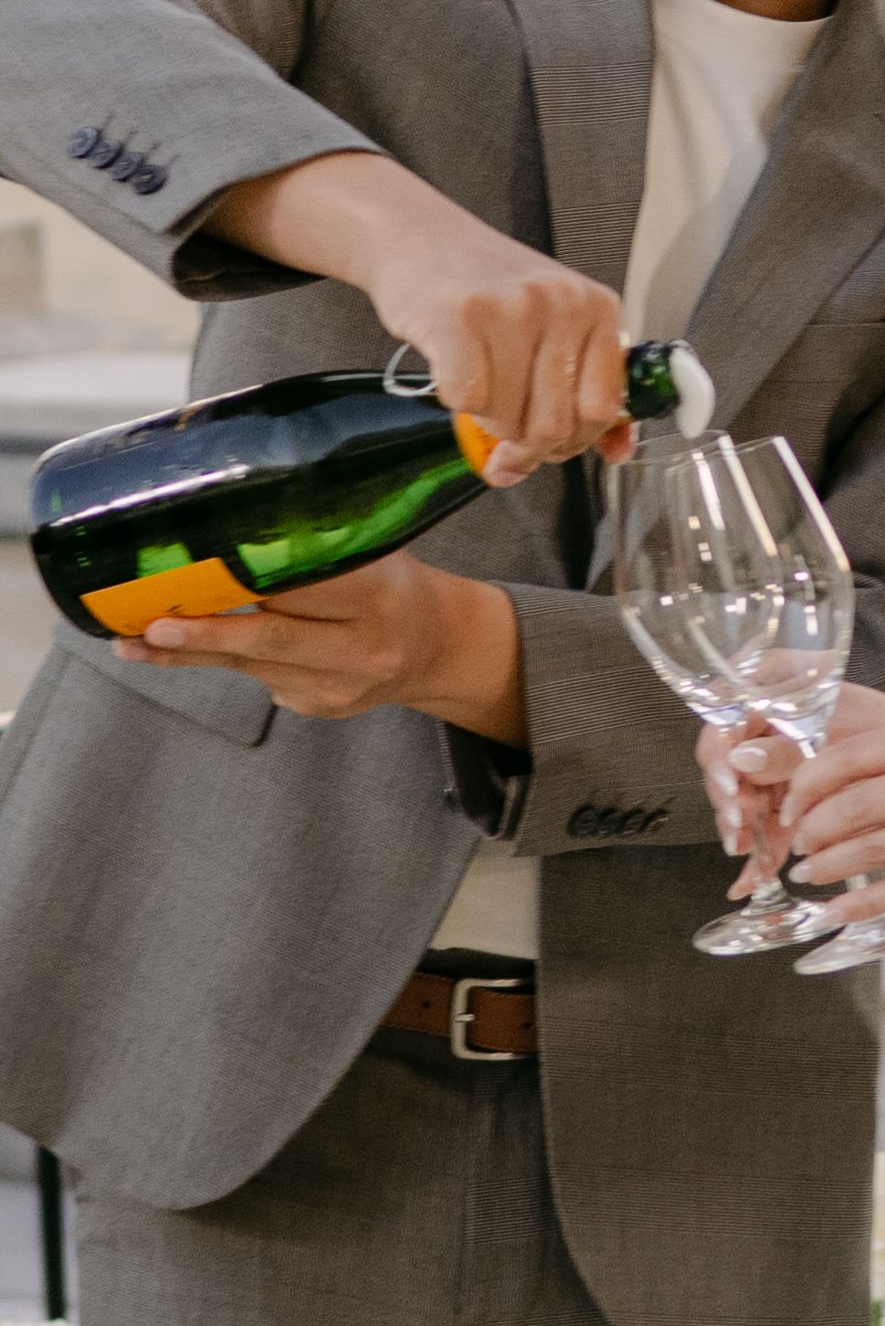 Person in a gray blazer pours sparkling wine from a green bottle with an orange label into two champagne glasses.