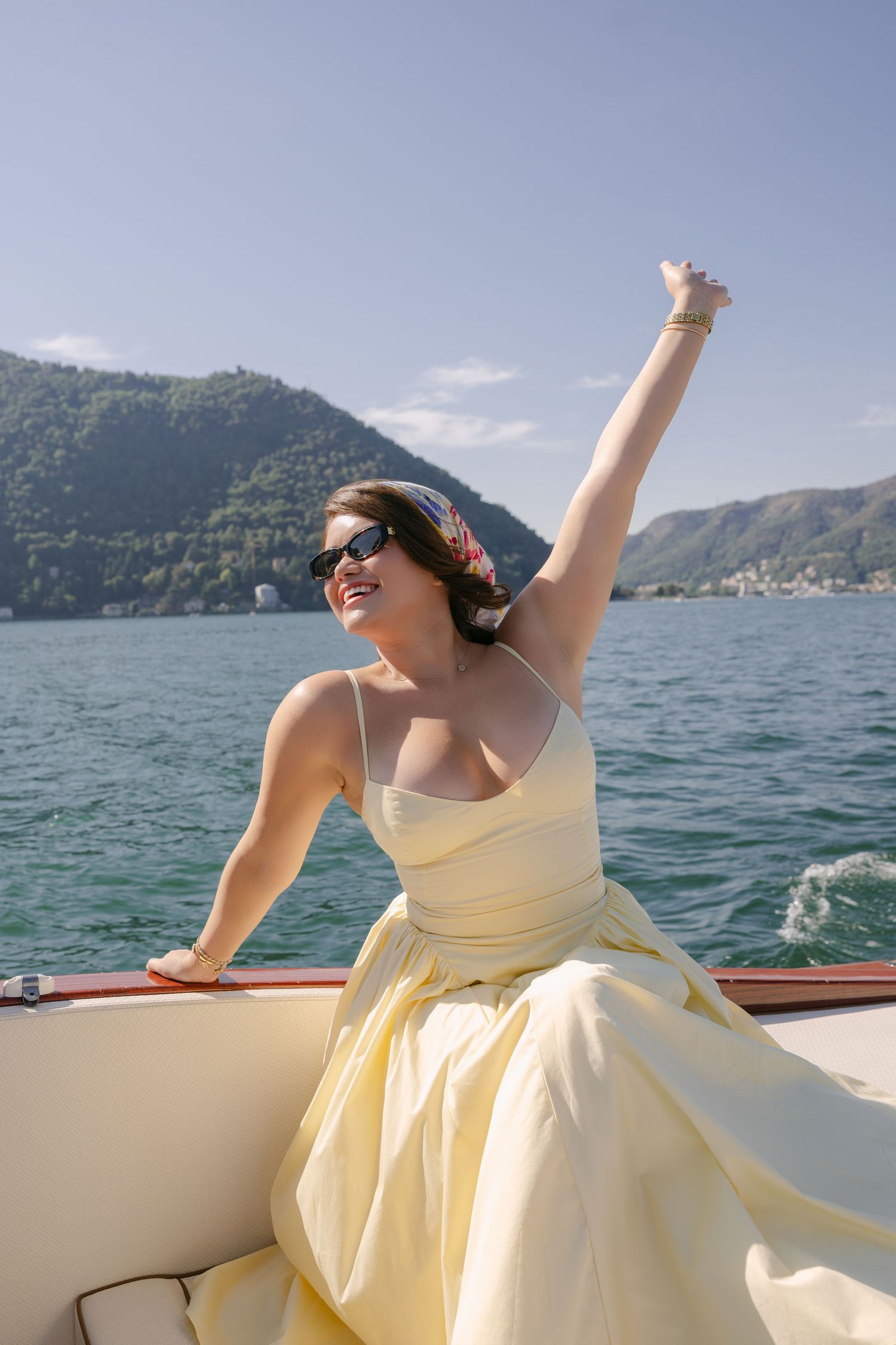 Woman in yellow dress celebrating on a boat with mountain lake views on a sunny day.