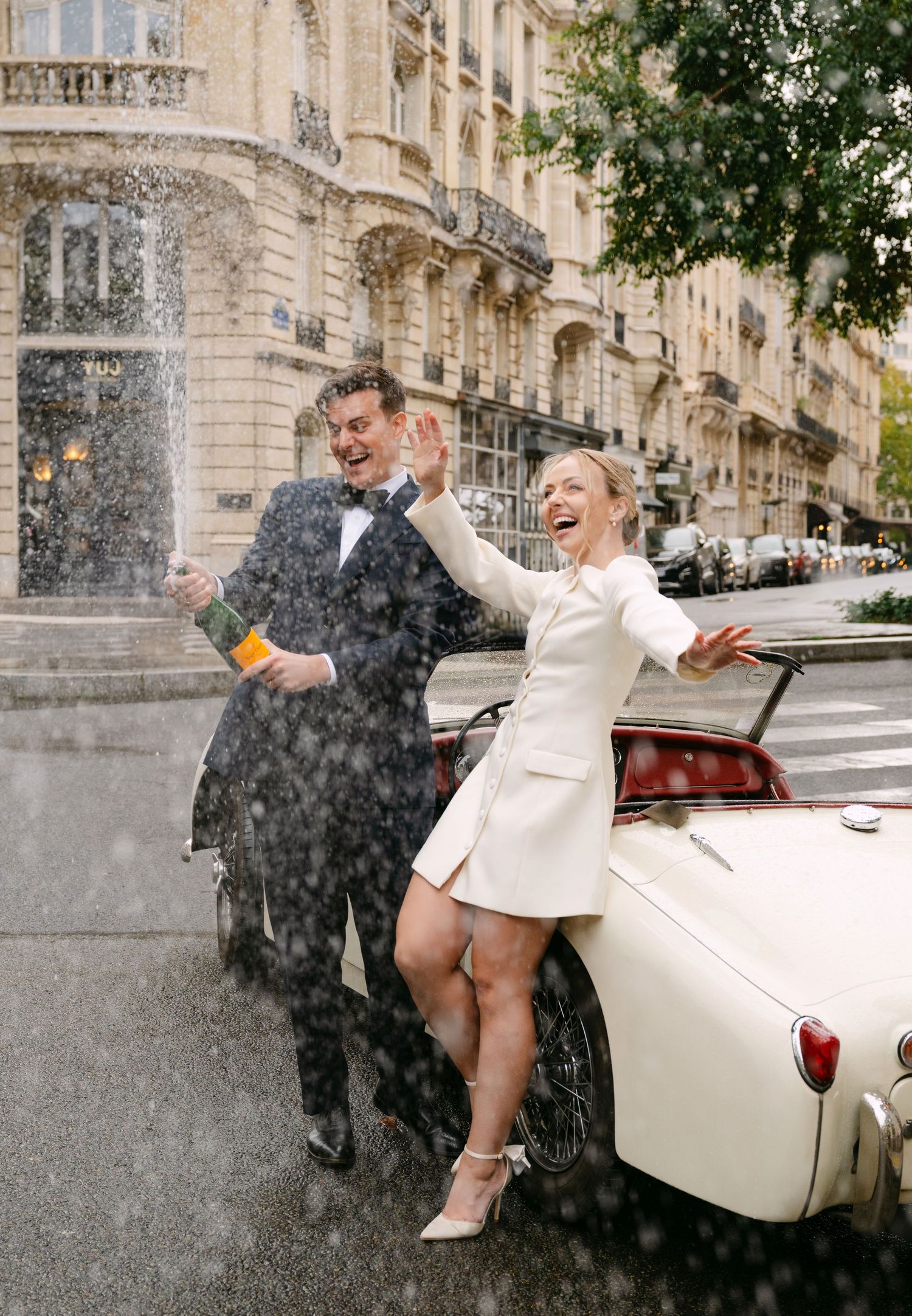 Joyful moment in the rain beside a classic white convertible car on a Parisian street.