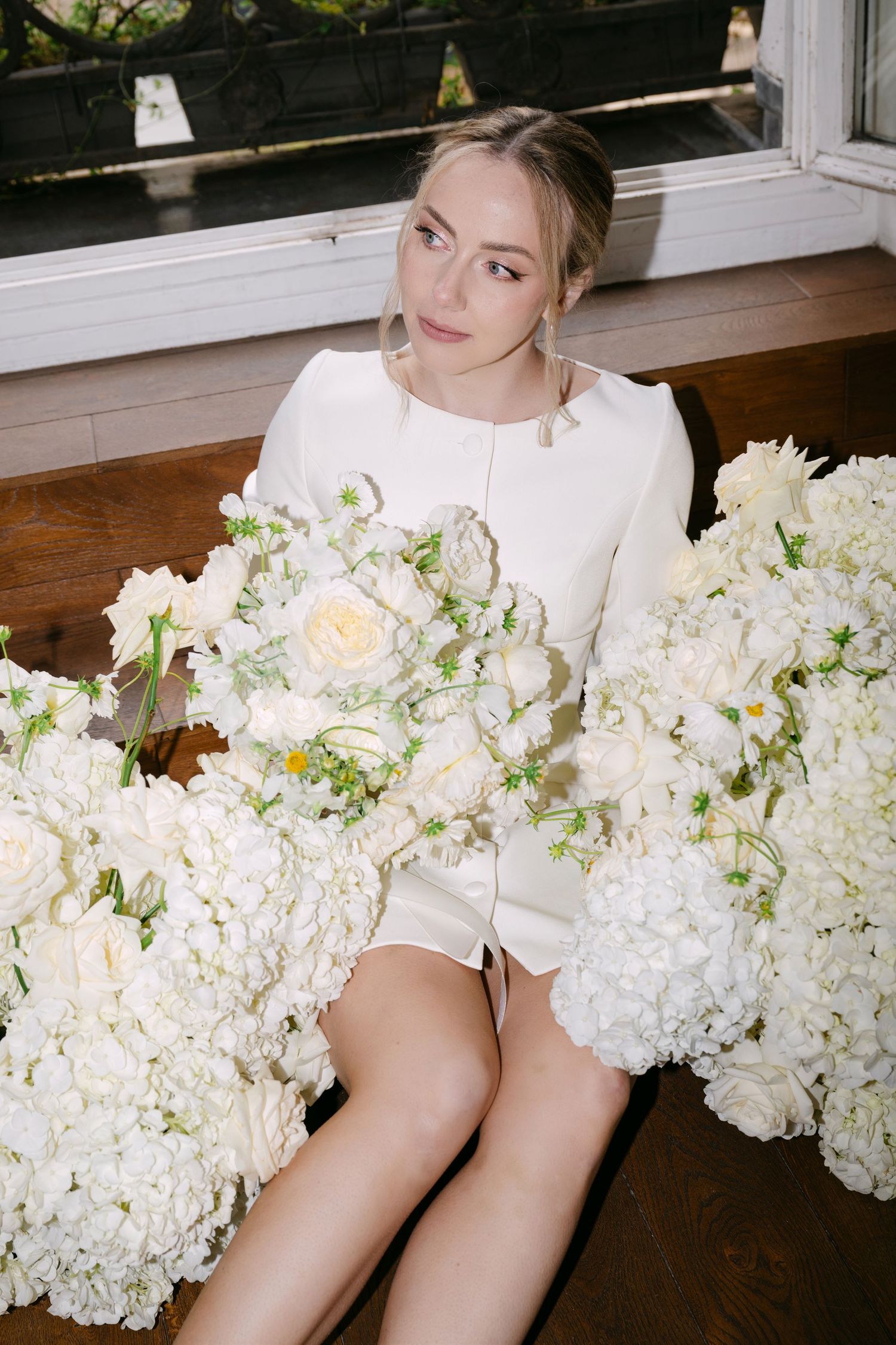 A person in a white sweater sits by a window surrounded by lush white hydrangeas and roses on a wooden surface.