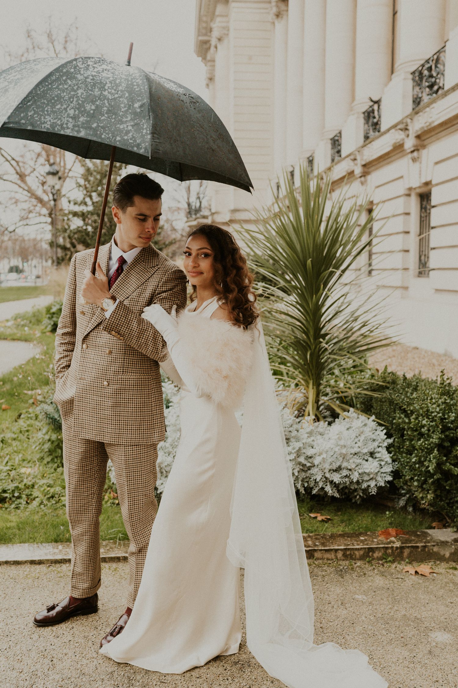 Vintage-styled wedding couple sharing an umbrella in front of a classic building with palm plants.