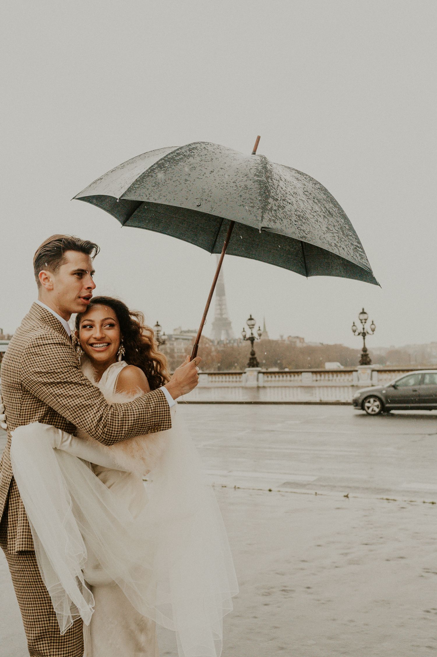Couple in wedding attire share intimate moment under umbrella on rainy Paris street.
