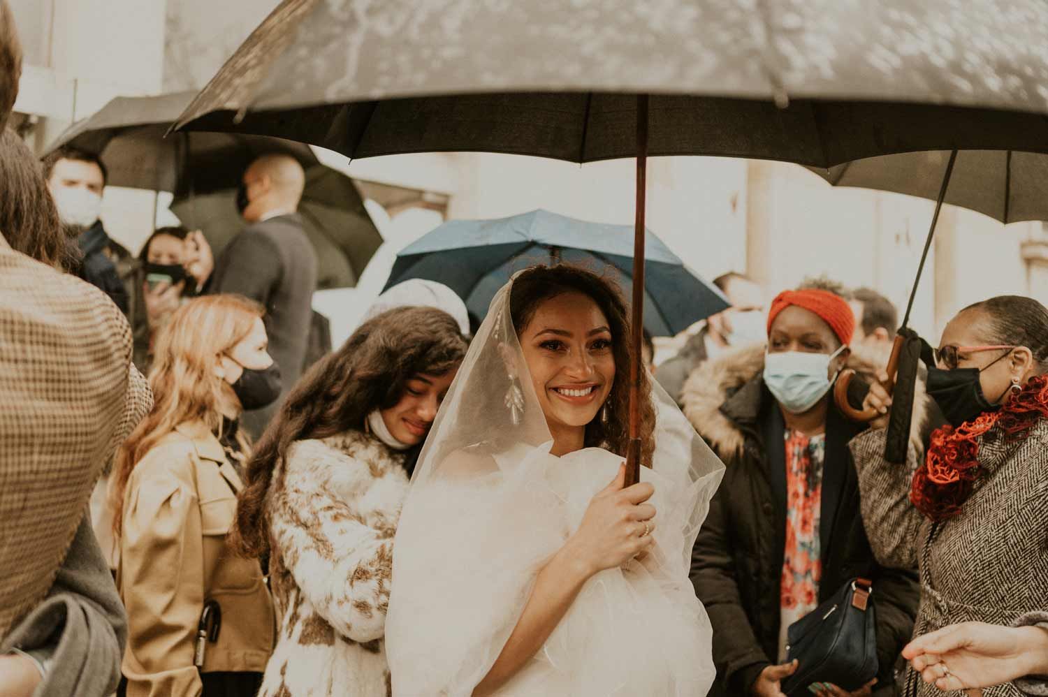A smiling bride under black umbrellas surrounded by wedding guests wearing face masks.