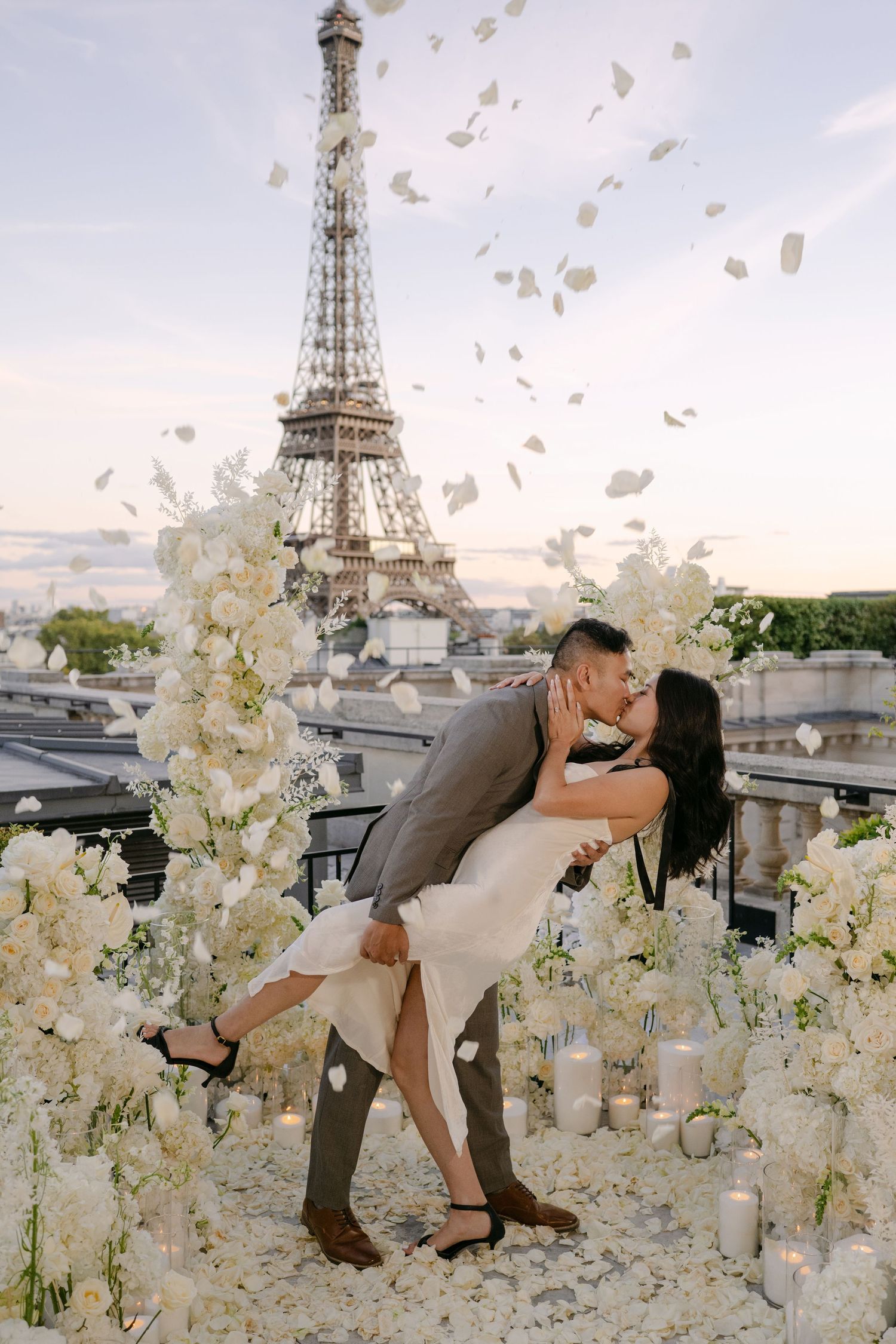 Couple kisses in a dramatic dip on a Paris rooftop surrounded by white flowers, candles and falling petals, with the Eiffel Tower behind.