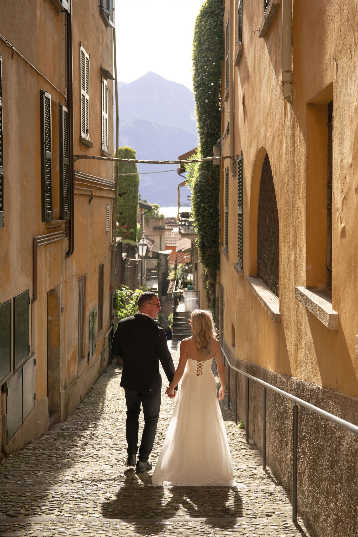 Bride and groom walking hand in hand down a cobblestone alley in an Italian village with mountains in the background.