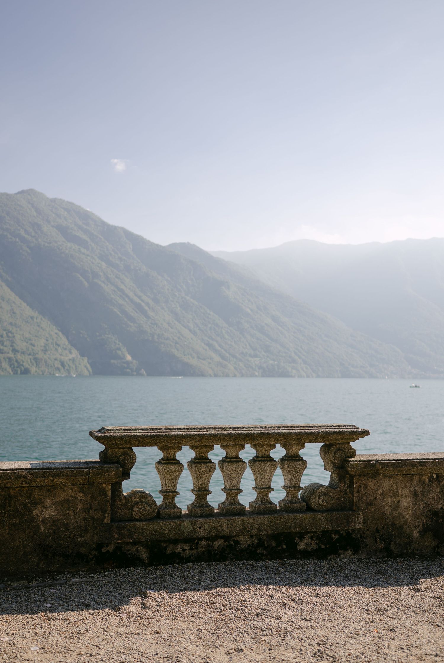 Stone balustrade overlooks serene Lake Como with misty mountain backdrop on a sunny day.