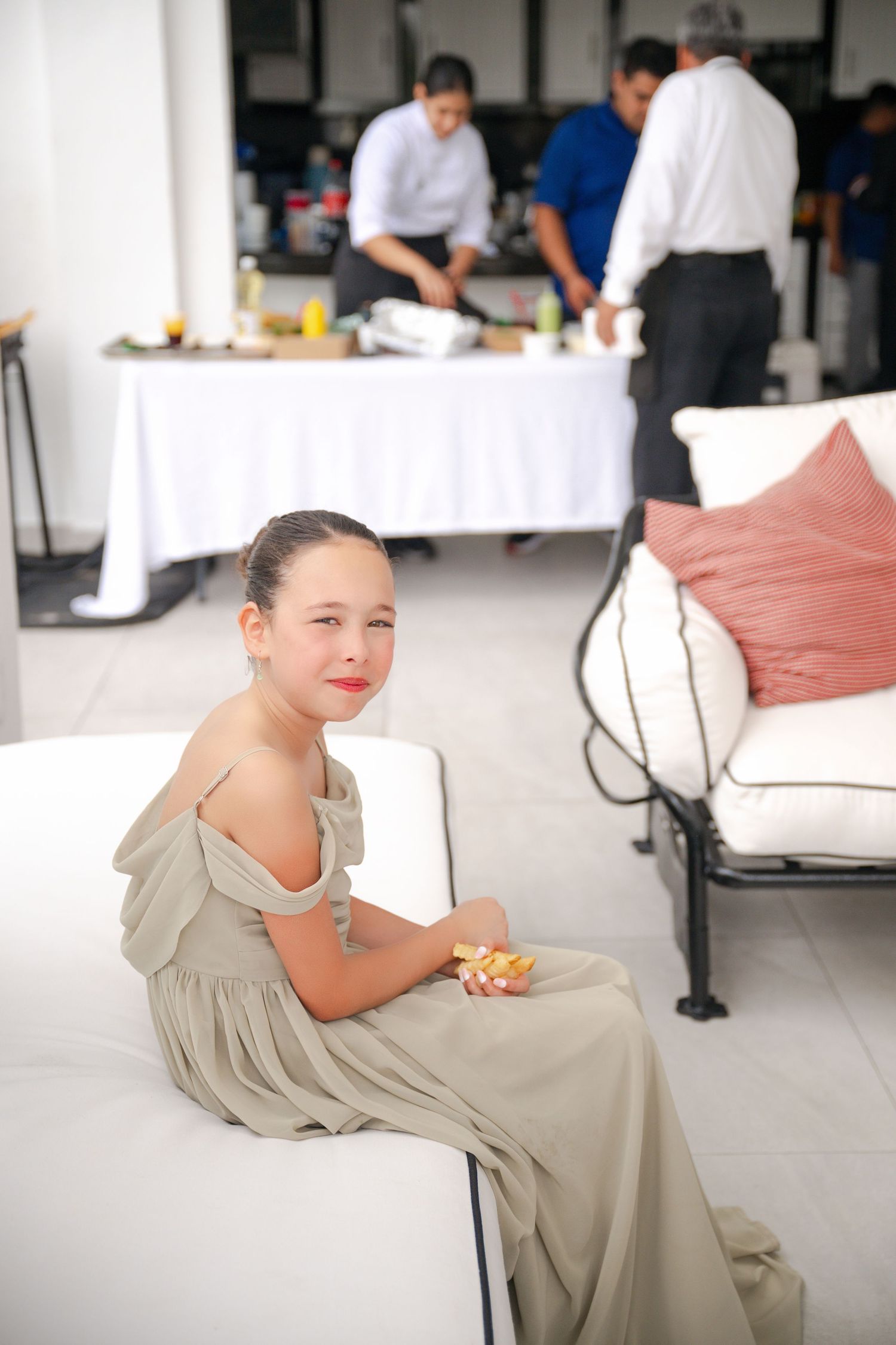Young girl in elegant gray dress sits on white sofa at an event, with catering staff preparing food table behind her.