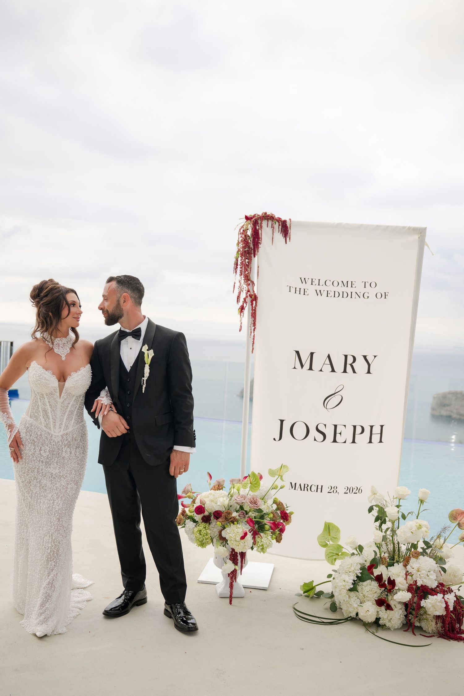 Bride and groom pose by elegant wedding welcome sign for Mary and Joseph, March 2025, with ocean backdrop.