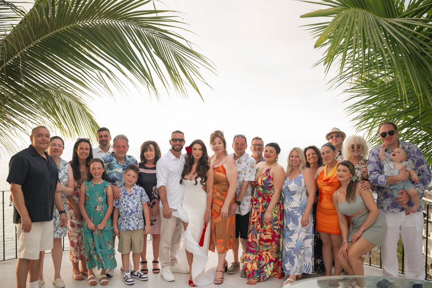 Large family group photo on tropical beach resort terrace, dressed in colorful summer attire, palm trees in background.