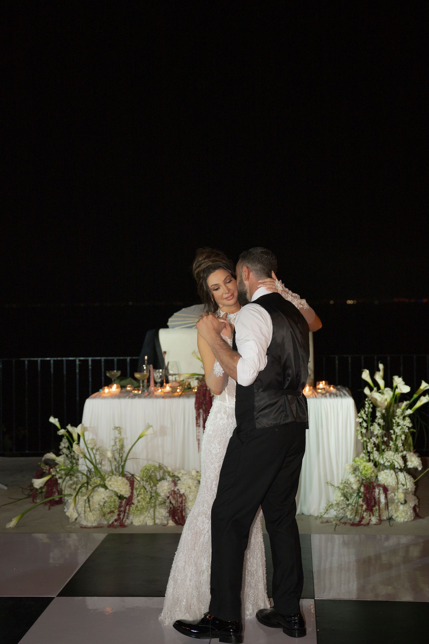 Bride and groom share their first dance outdoors at night, with candlelit floral arrangements and wedding cake in background.