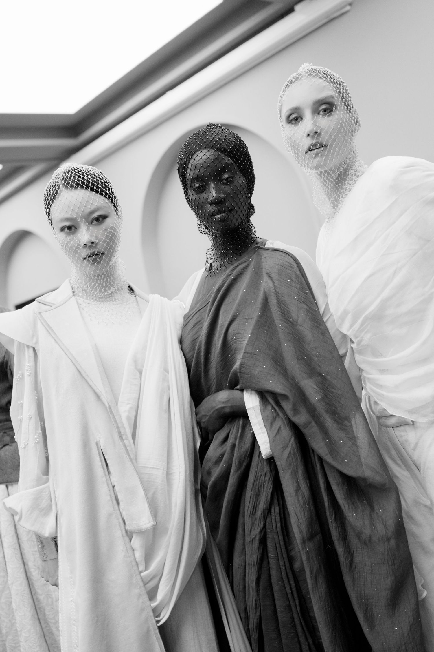 Three models in draped white and dark garments with mesh veils pose together in a black and white fashion editorial.