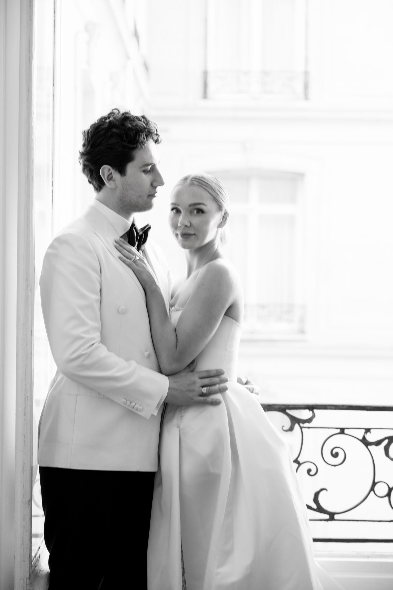Black and white photo of bride and groom embracing by a Parisian balcony window in wedding attire.