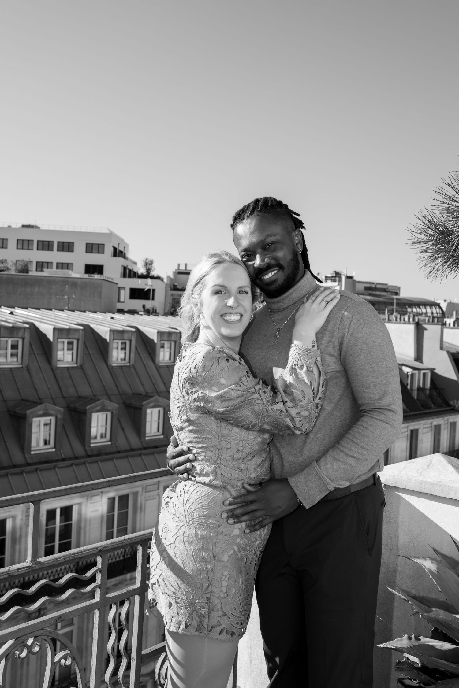 Happy interracial couple embracing on a Parisian rooftop terrace, smiling in black and white photo.