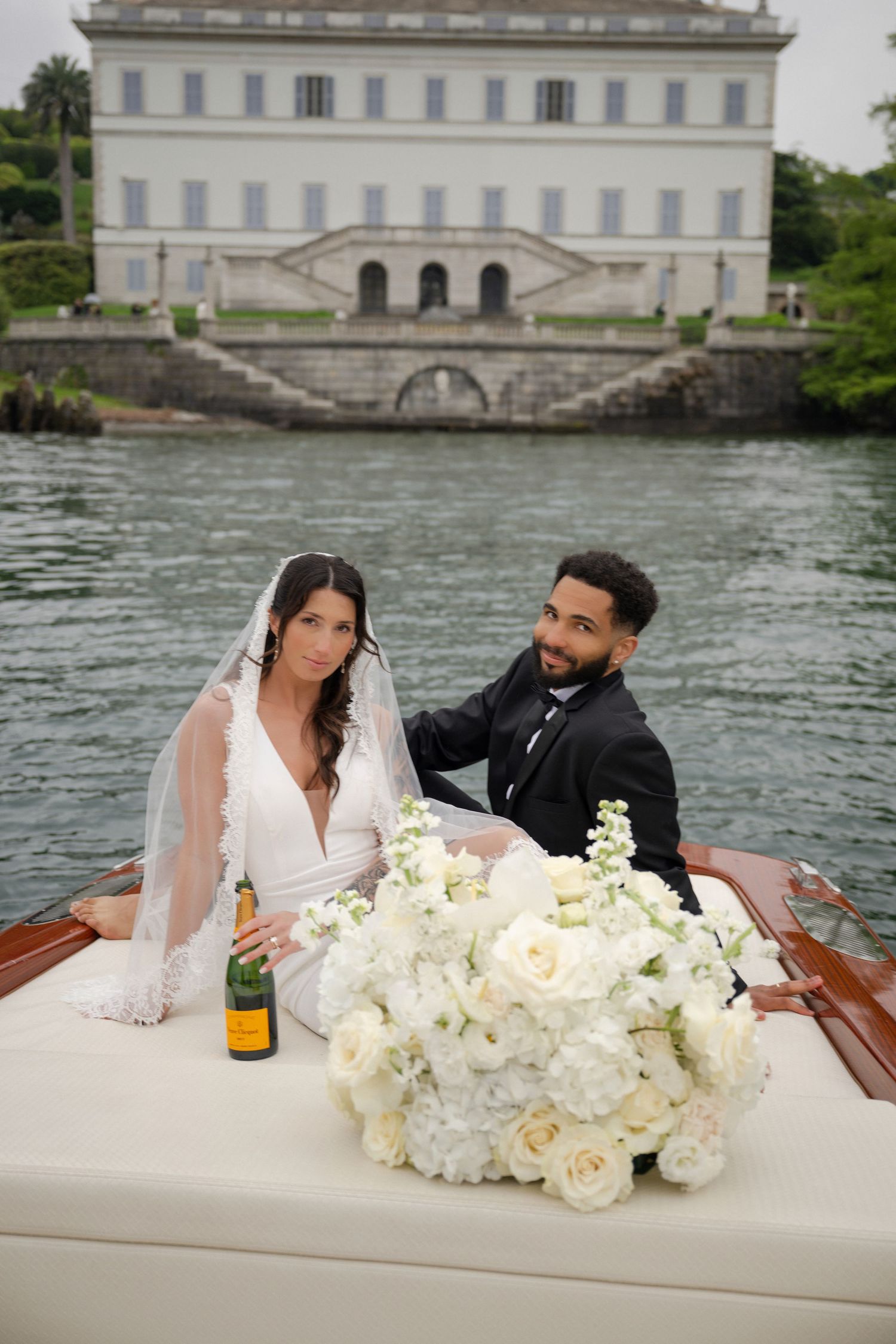 Bride and groom sitting on a classic wooden boat with white floral bouquet and champagne before a grand villa.