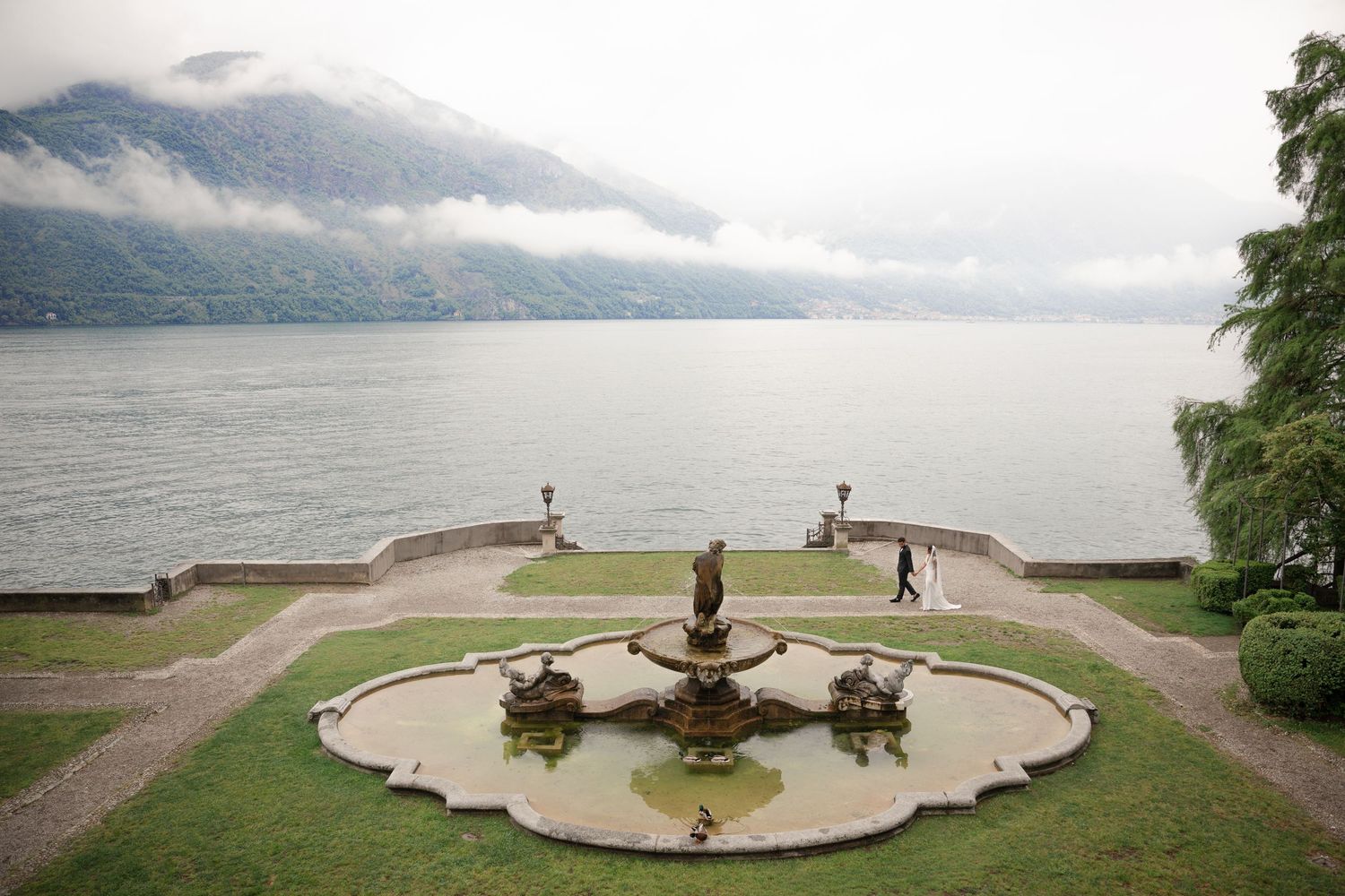 Elegant Italian villa garden with ornate fountain overlooking misty lake Como with mountain views.