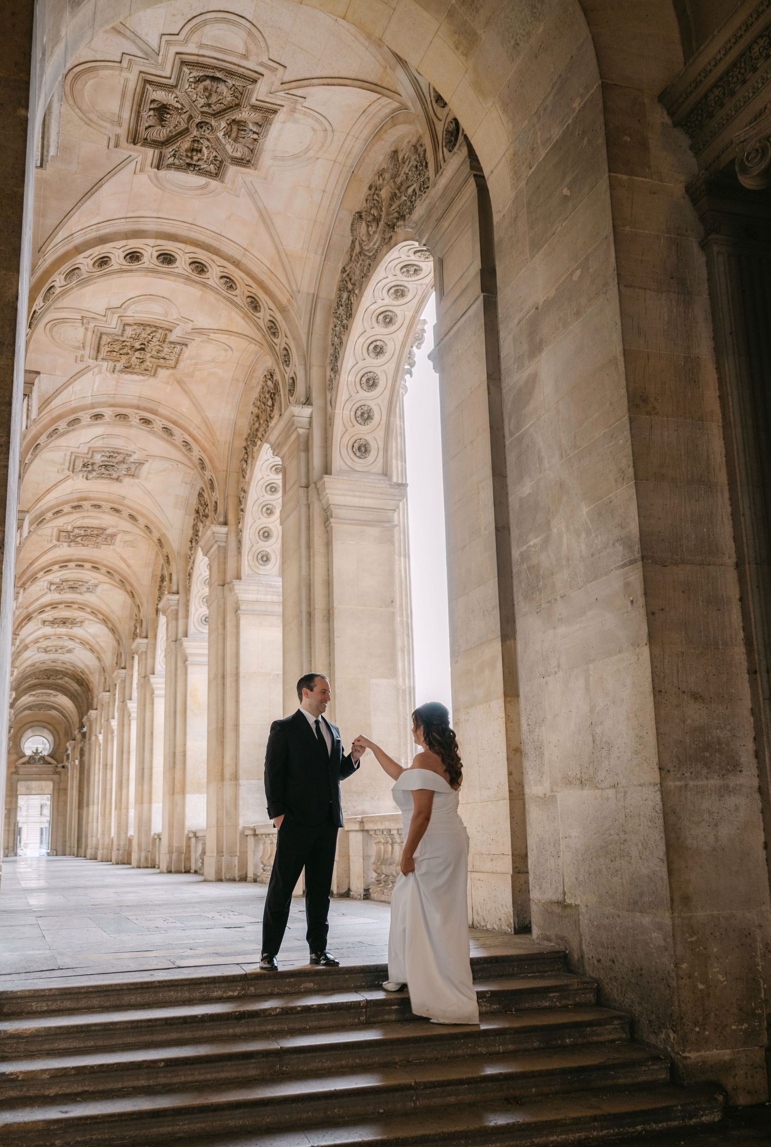 Couple in wedding attire dancing under ornate arched colonnade of historic European building.
