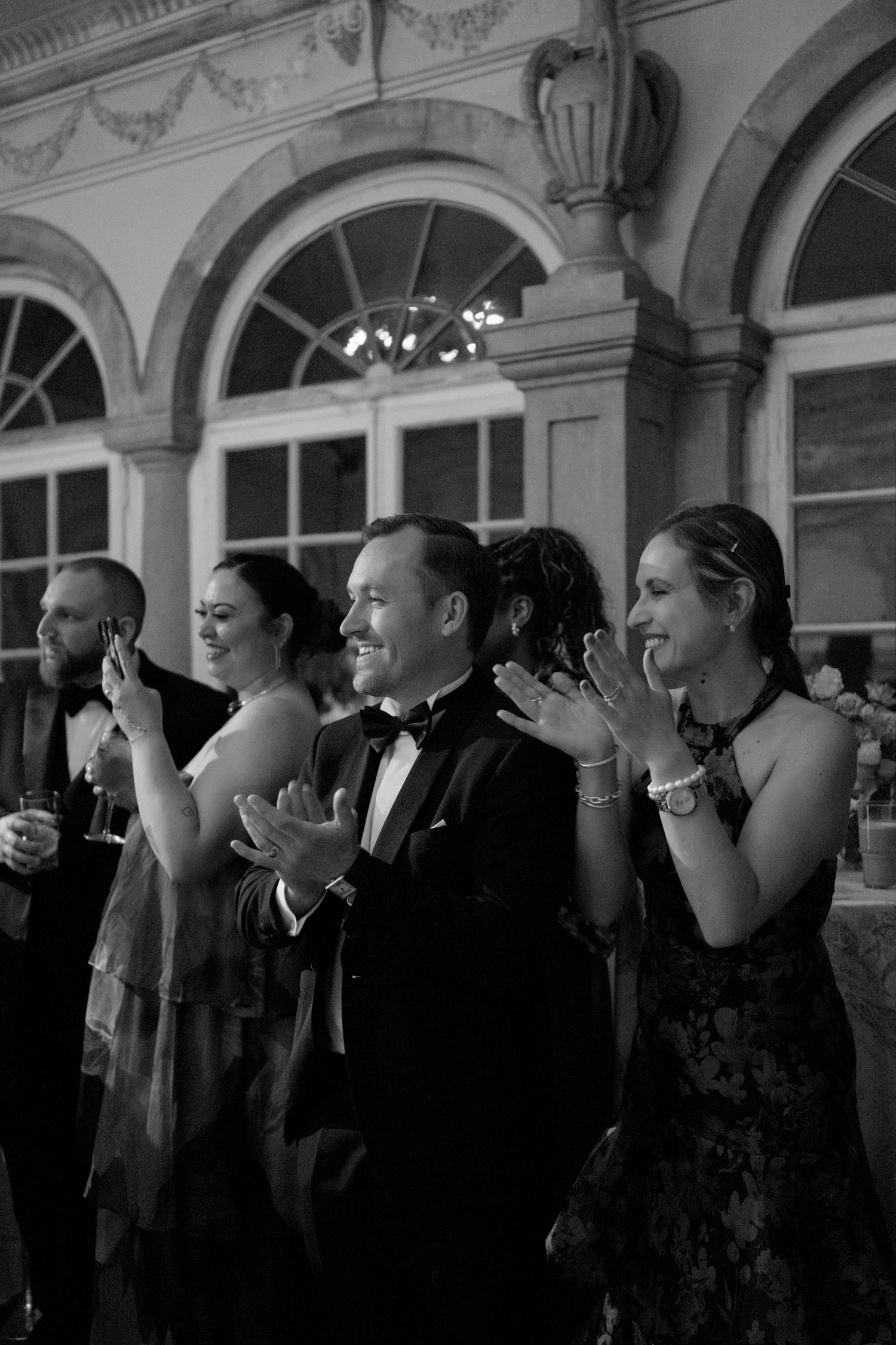Elegantly dressed guests clapping and smiling at a formal black-tie event in a grand ballroom.