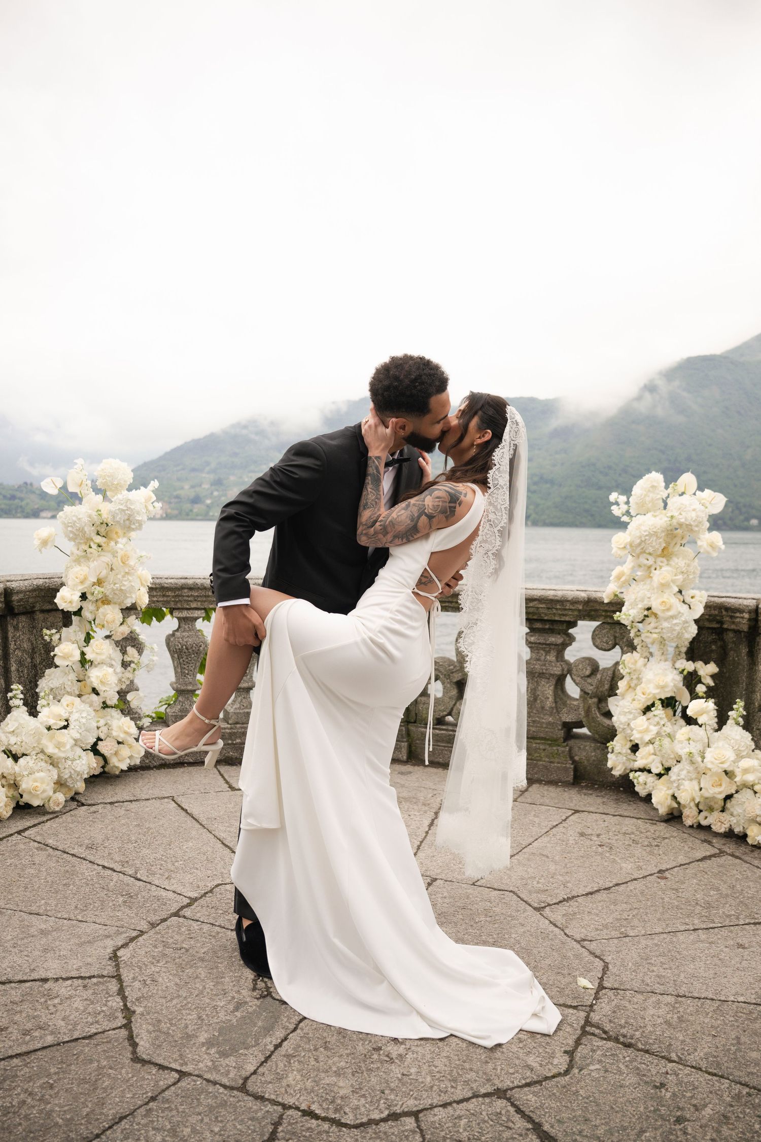Bride and groom share a romantic dip kiss on a lakeside terrace adorned with white floral arrangements on their wedding day.